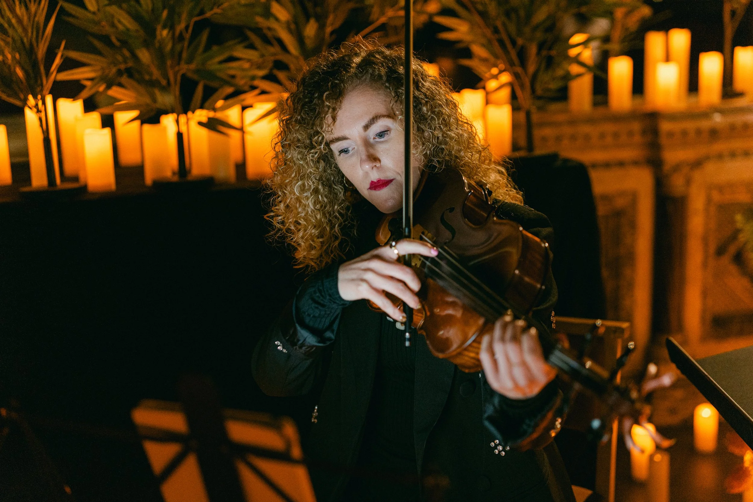 A woman with curly red hair playing a violin in a dimly lit room with warm candlelight in the background.