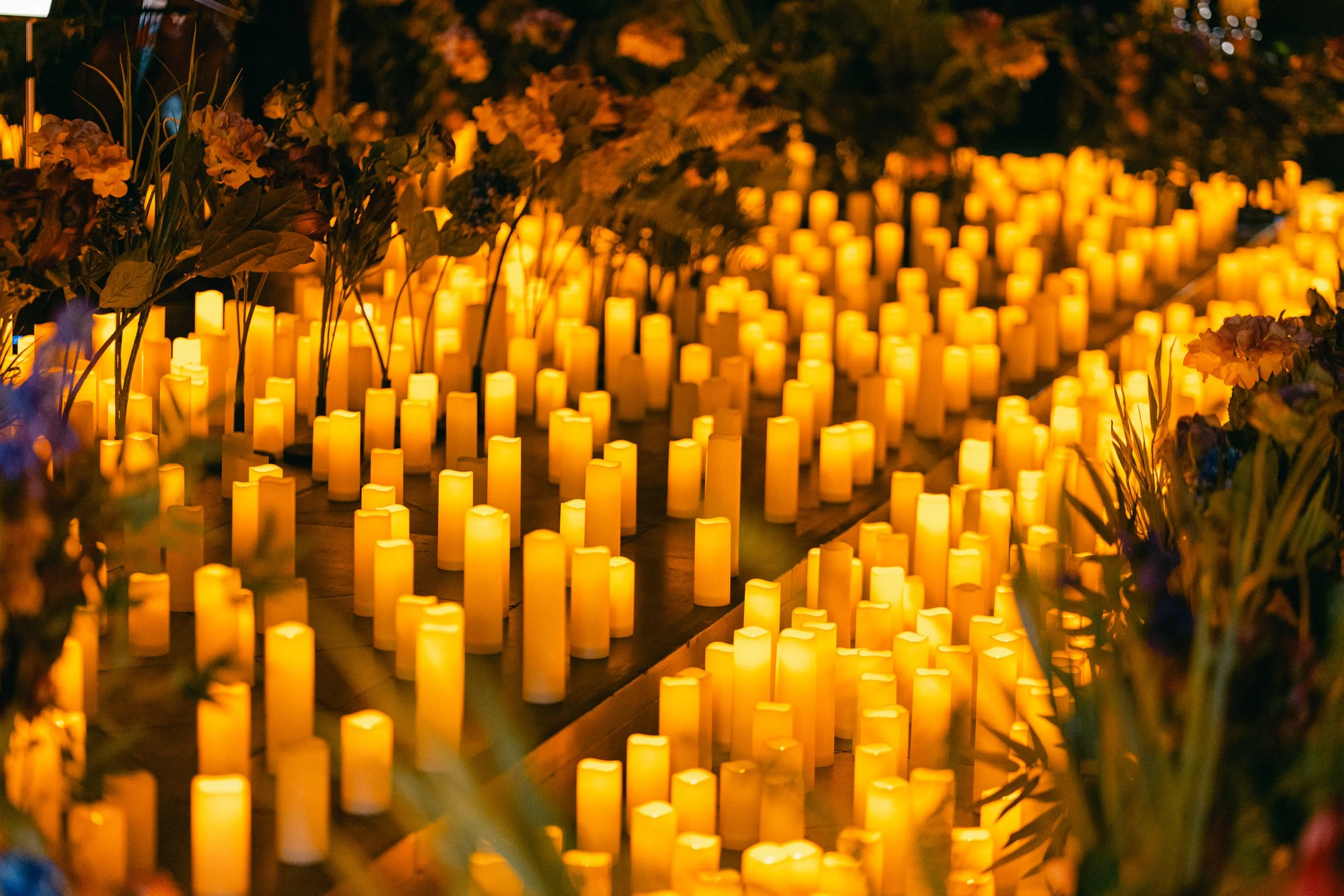 Numerous yellow candles arranged on wooden surfaces, surrounded by flowers and greenery, creating a warm, glowing scene.