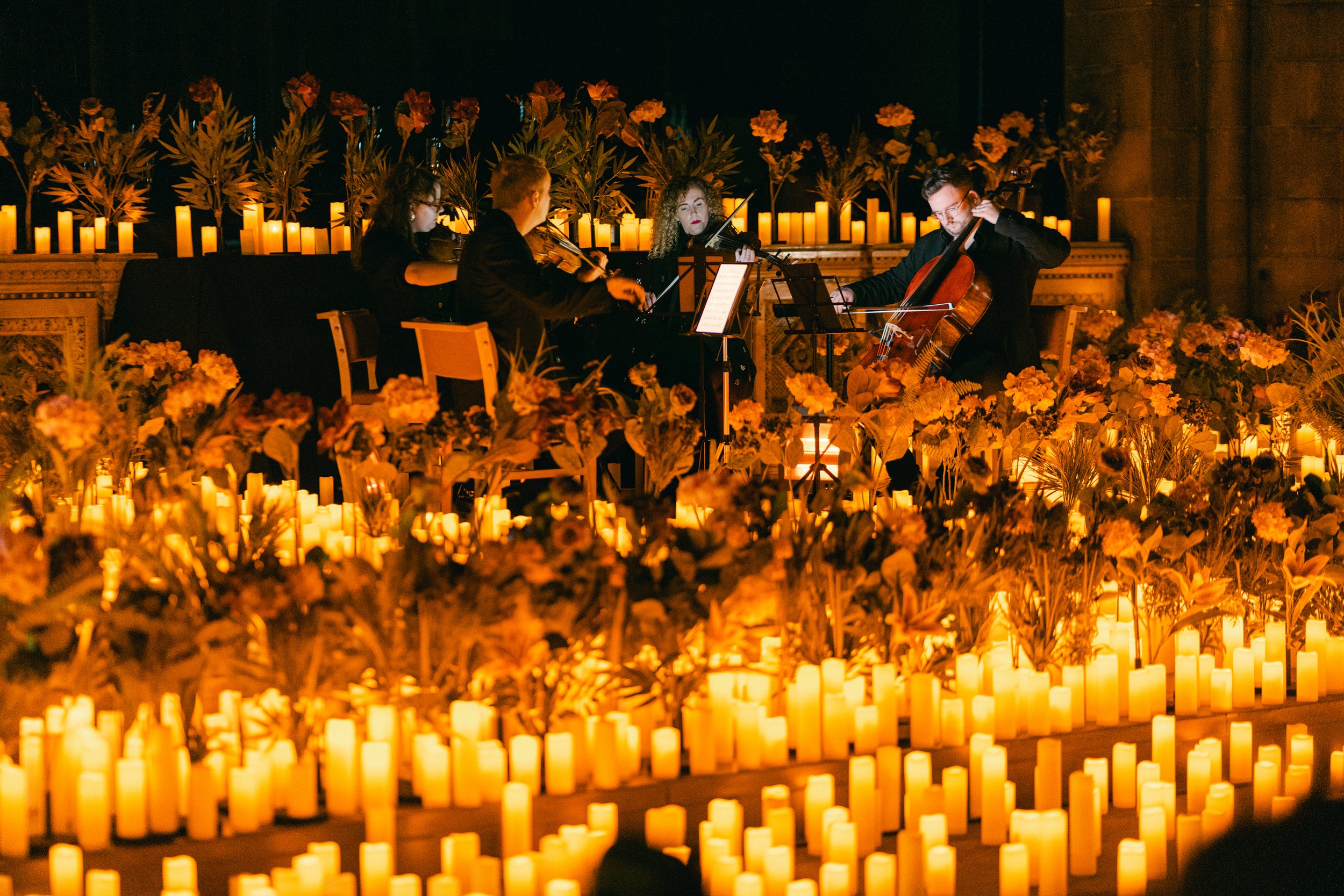 A string of four musicians performing a classical concert, playing violins and a cello, surrounded by candles and flowers in a dimly lit setting.