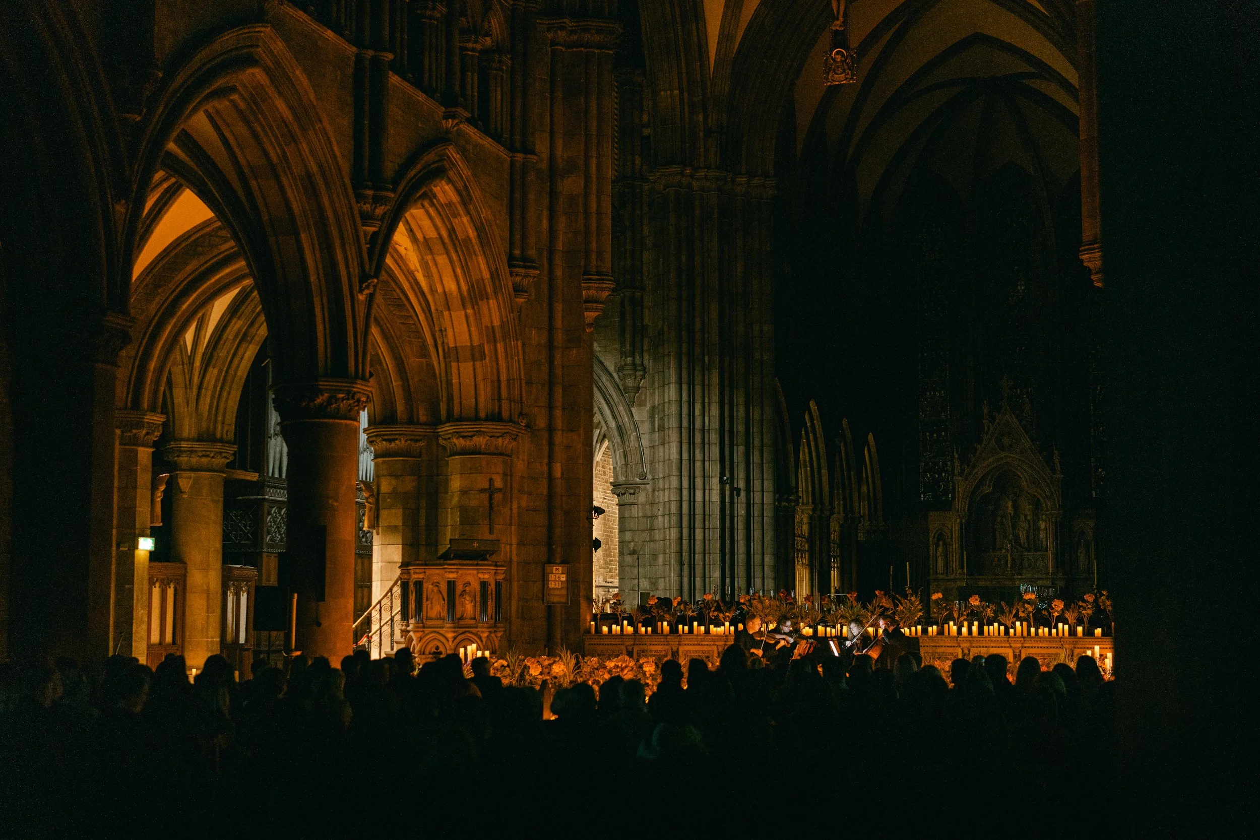 A nighttime scene inside a dimly lit gothic cathedral with pointed arches and ornate architecture. The interior features a large congregation of people seated in dark silhouettes, facing a decorated altar illuminated by numerous candles and floral ar