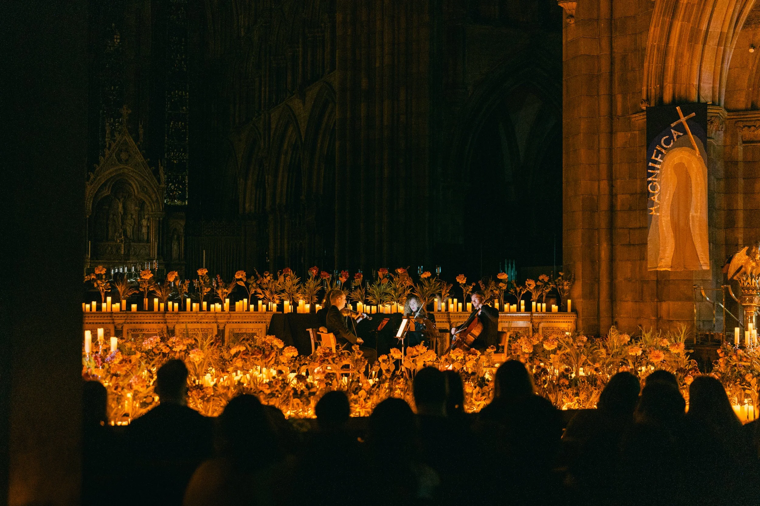 Musicians playing string instruments on a candlelit stage inside a large, Gothic-style cathedral with artwork and Gothic arches.
