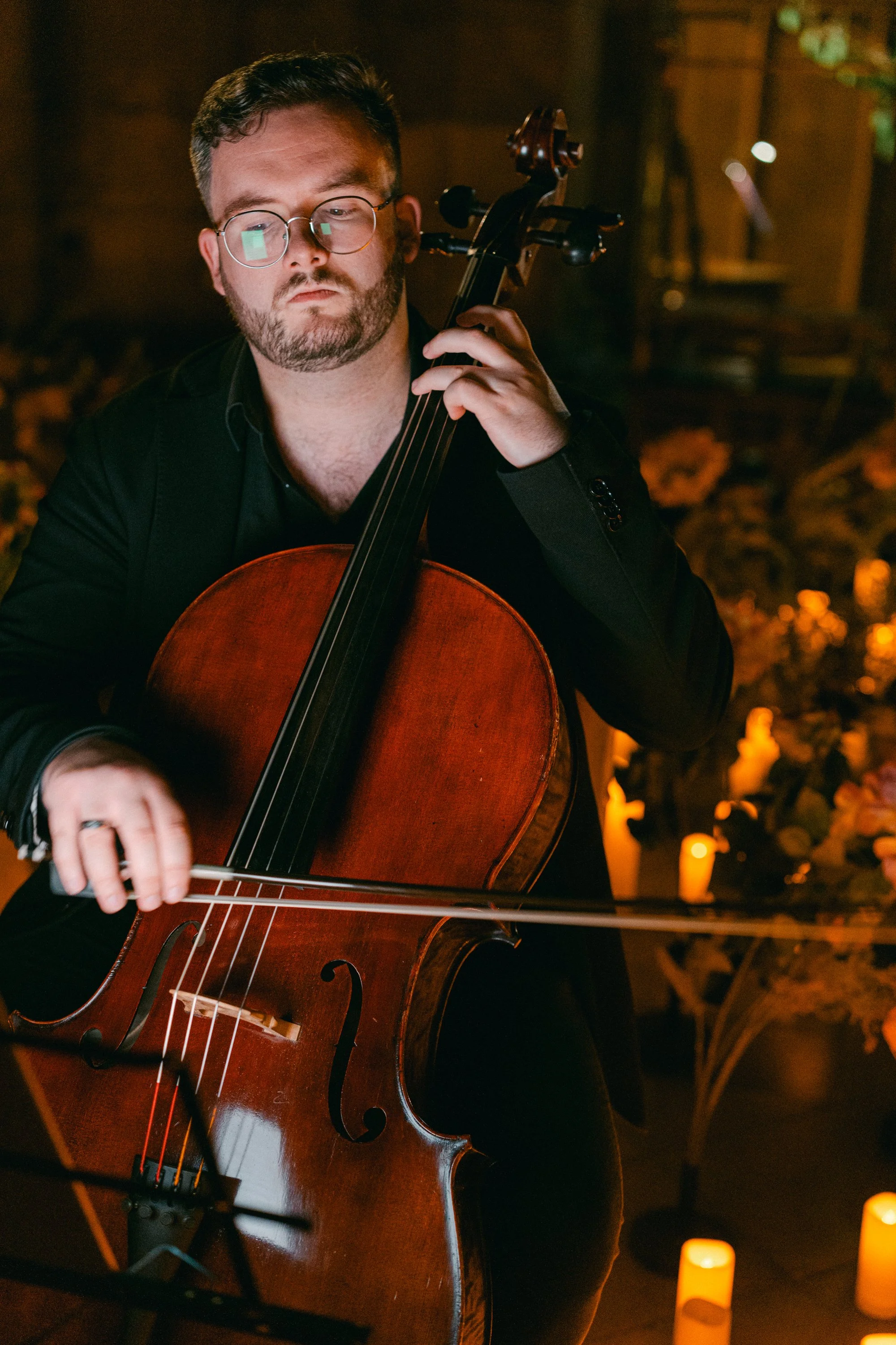 A man with glasses and a beard playing a cello in a warmly lit environment with candles and floral decorations.