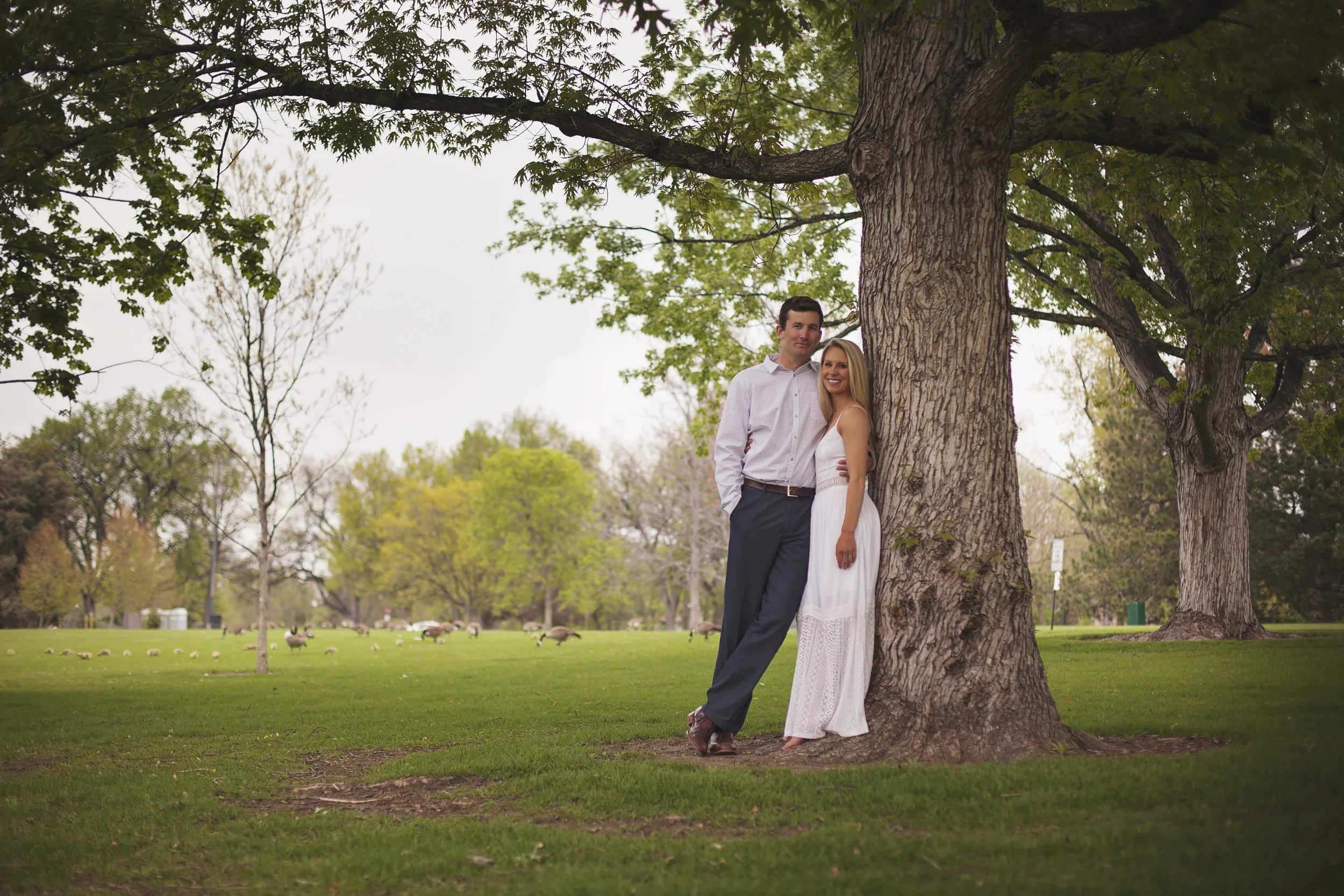 Couple leaning on tree with green grass around them during engagement photo session