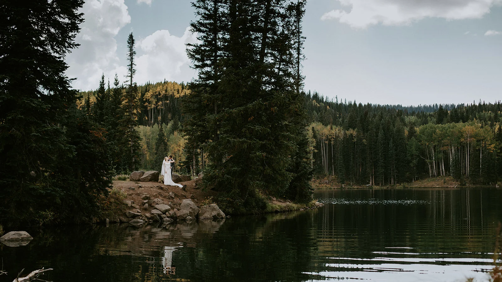 Wide wedding photo of Sunset Lake where bride and groom embrace and fall colors at Mesa Lakes Lodge