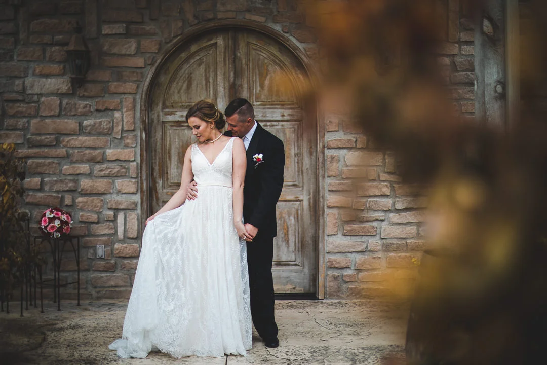 Bride holding dress with groom kissing her shoulder in front of arched winery door