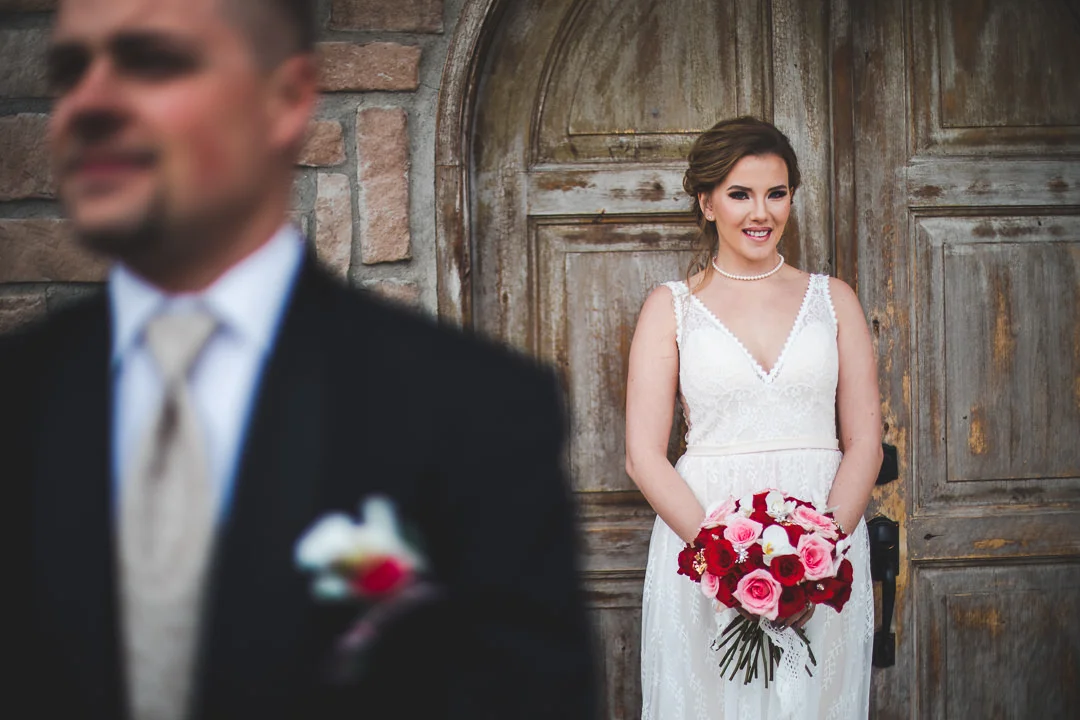 Groom in foreground out of focus with bride in background in front of wooden door in focus
