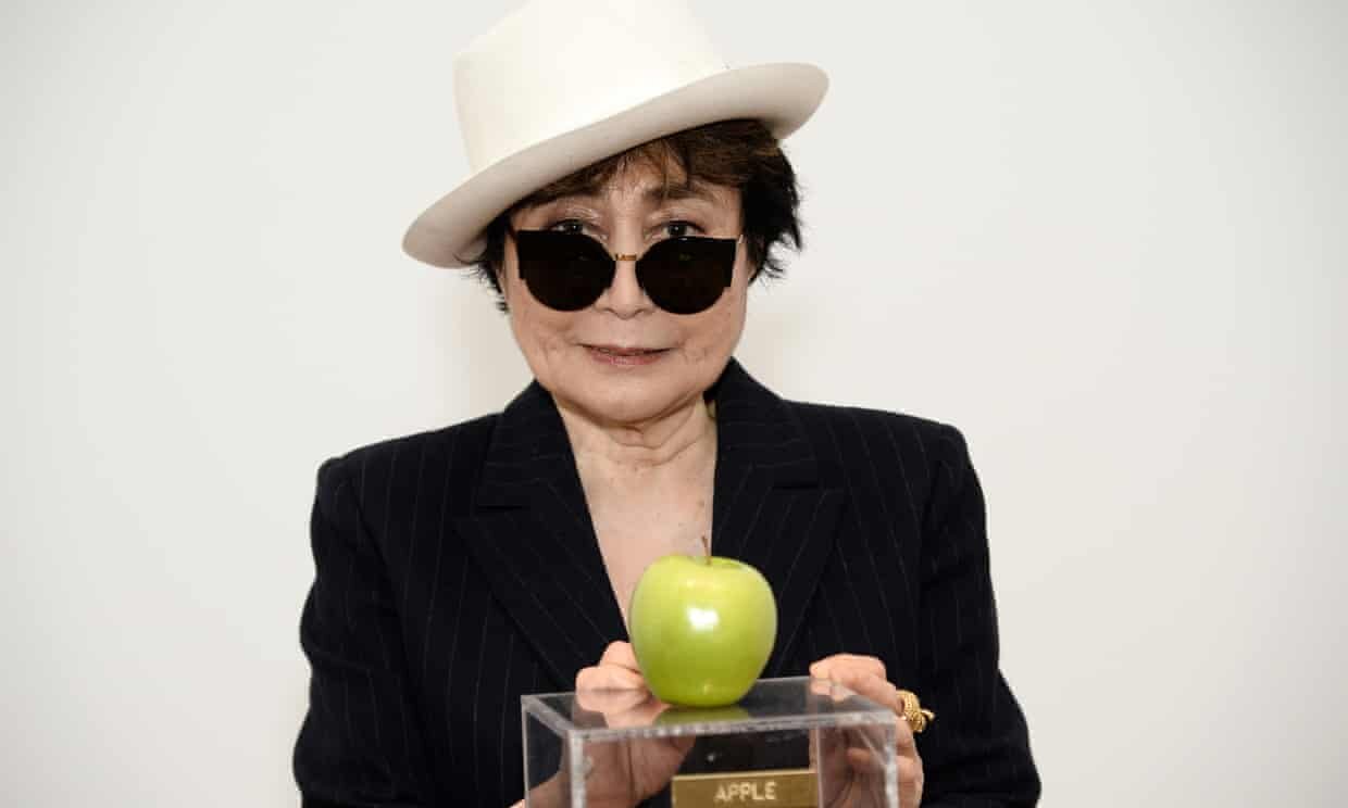 A woman wearing a white hat, black sunglasses, and a dark pinstripe blazer, holding a green apple on a clear display stand with a gold plaque that reads 'APPLE'.