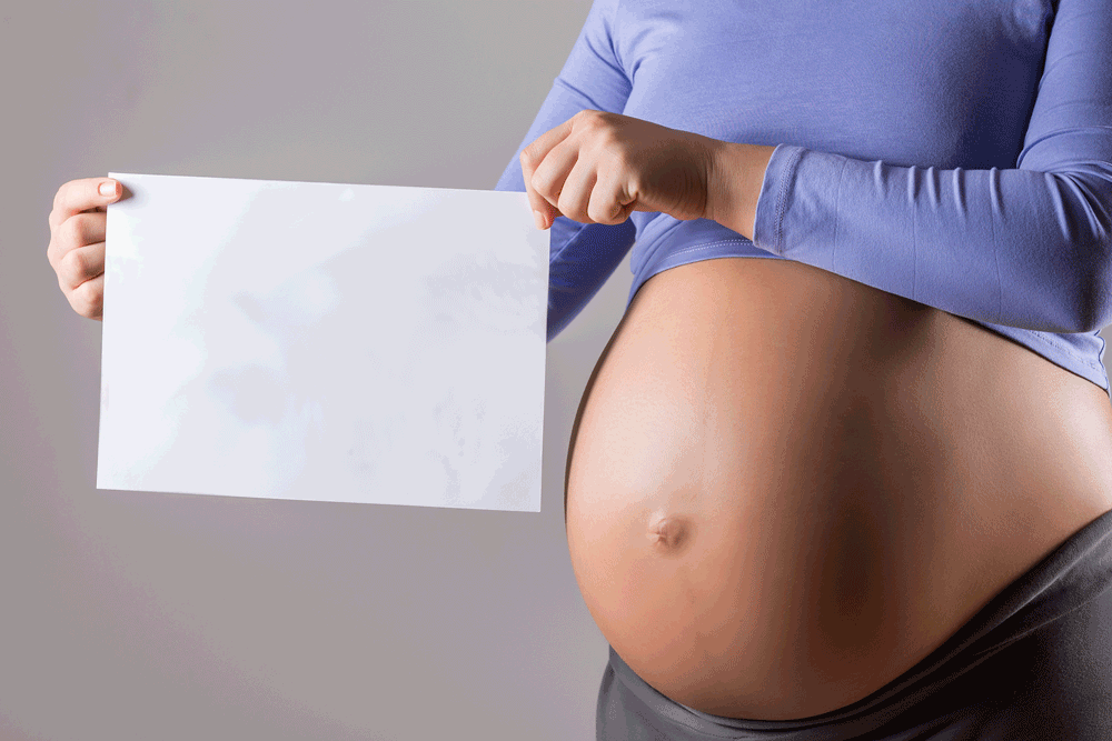 Pregnant woman holding a blank white piece of paper, wearing a blue shirt rolled up to reveal belly.