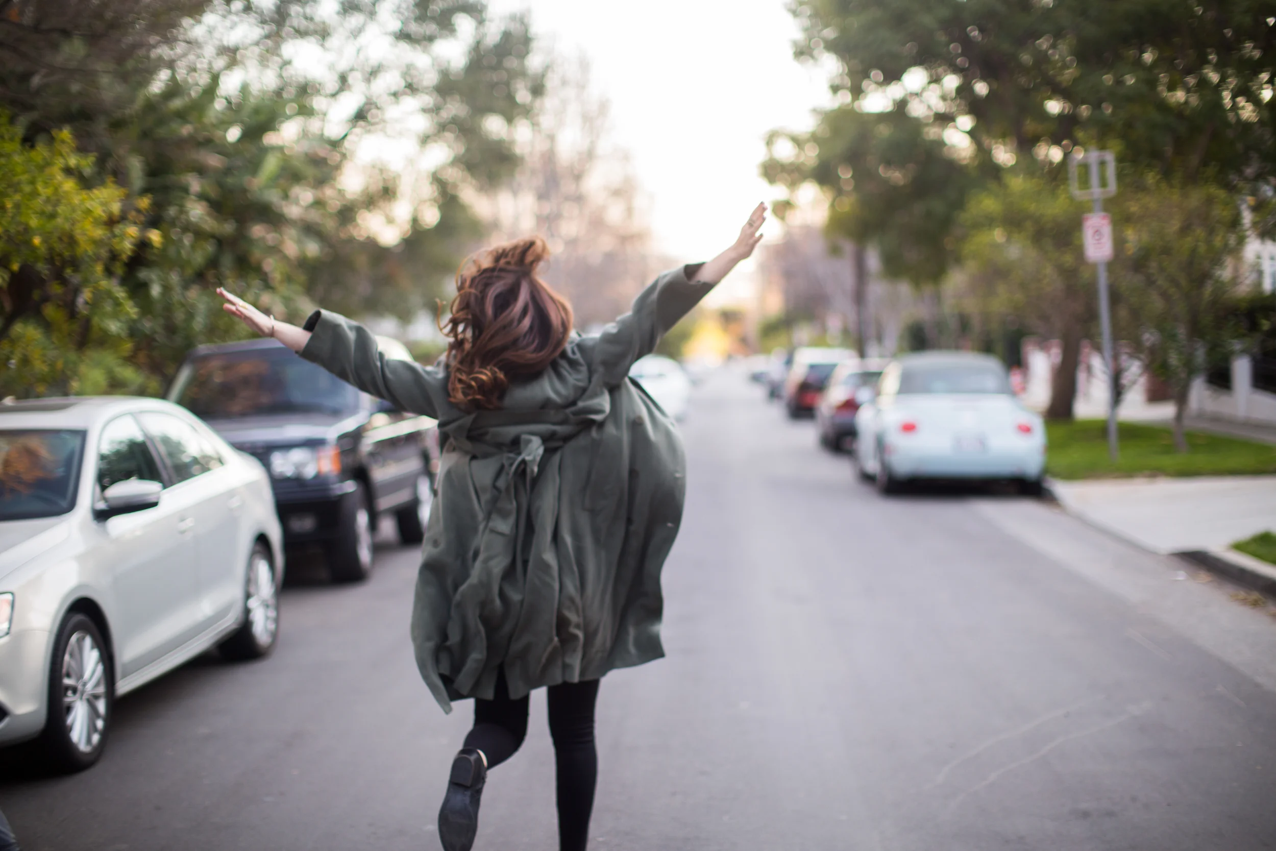 Woman in an olive jacket walking down a tree-lined street with her arms raised, lifestyle photography (Copy)