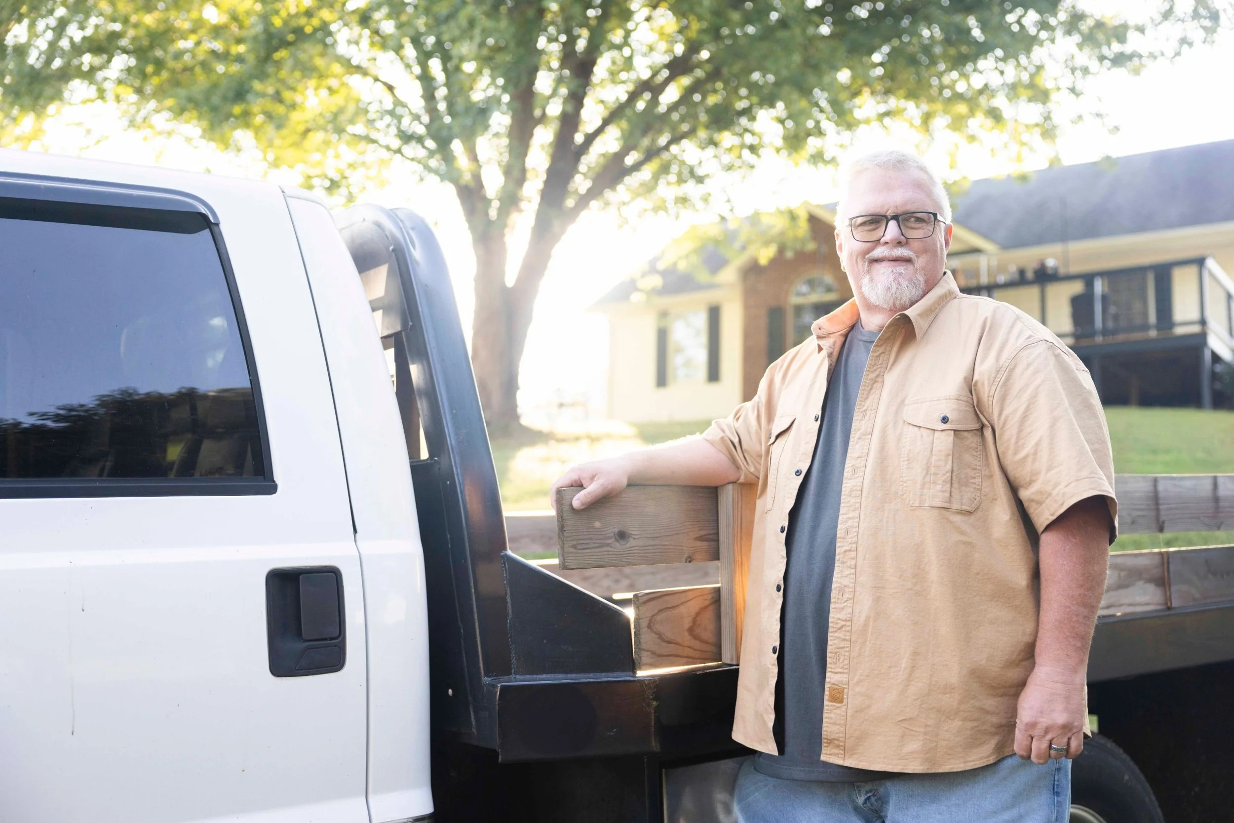 Pharma patient testimonial photography: man in a tan work shirt smiling beside a flatbed truck at his home in Tennessee
