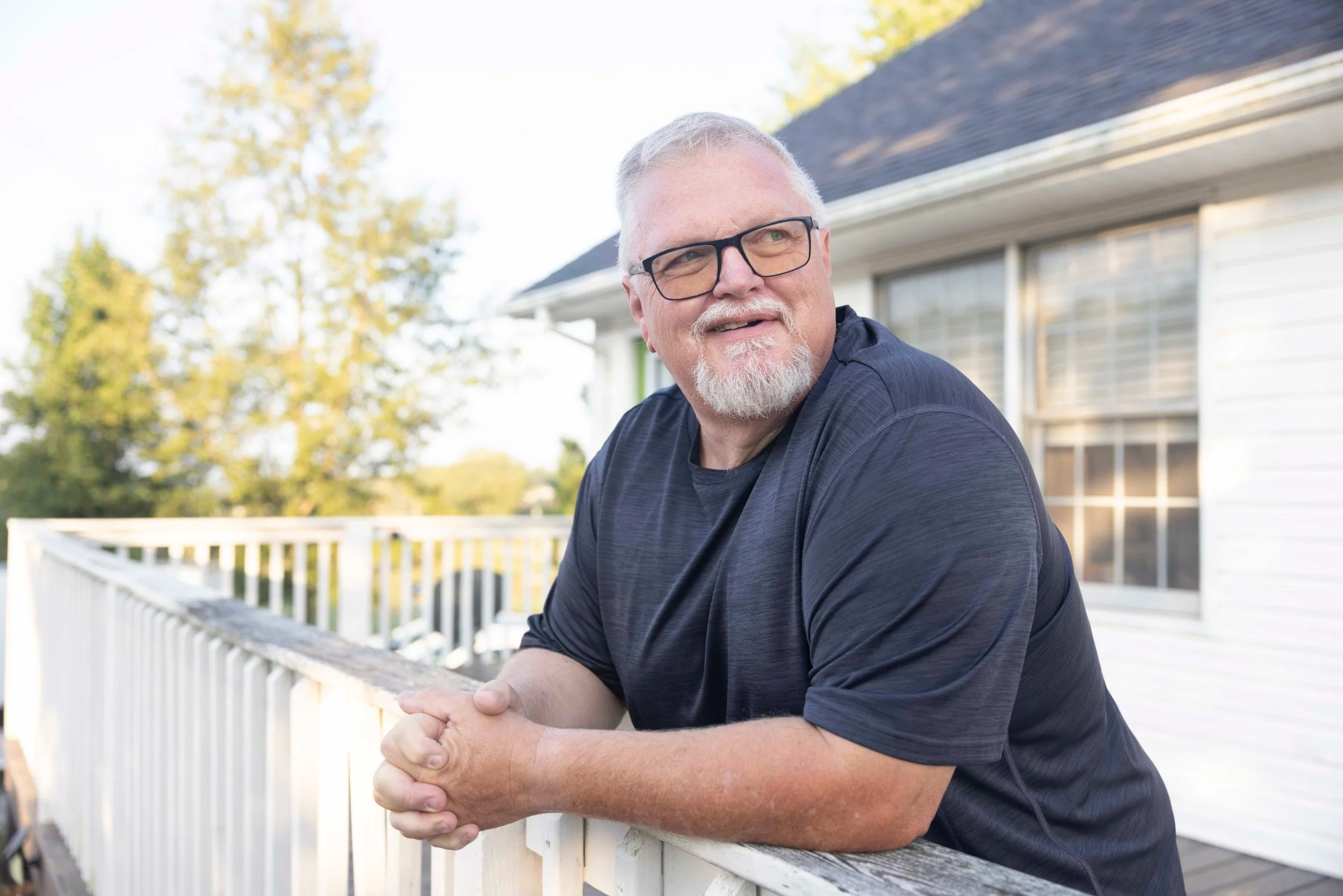 Healthcare patient photography: man in glasses leaning on a white deck railing, looking off-camera and smiling