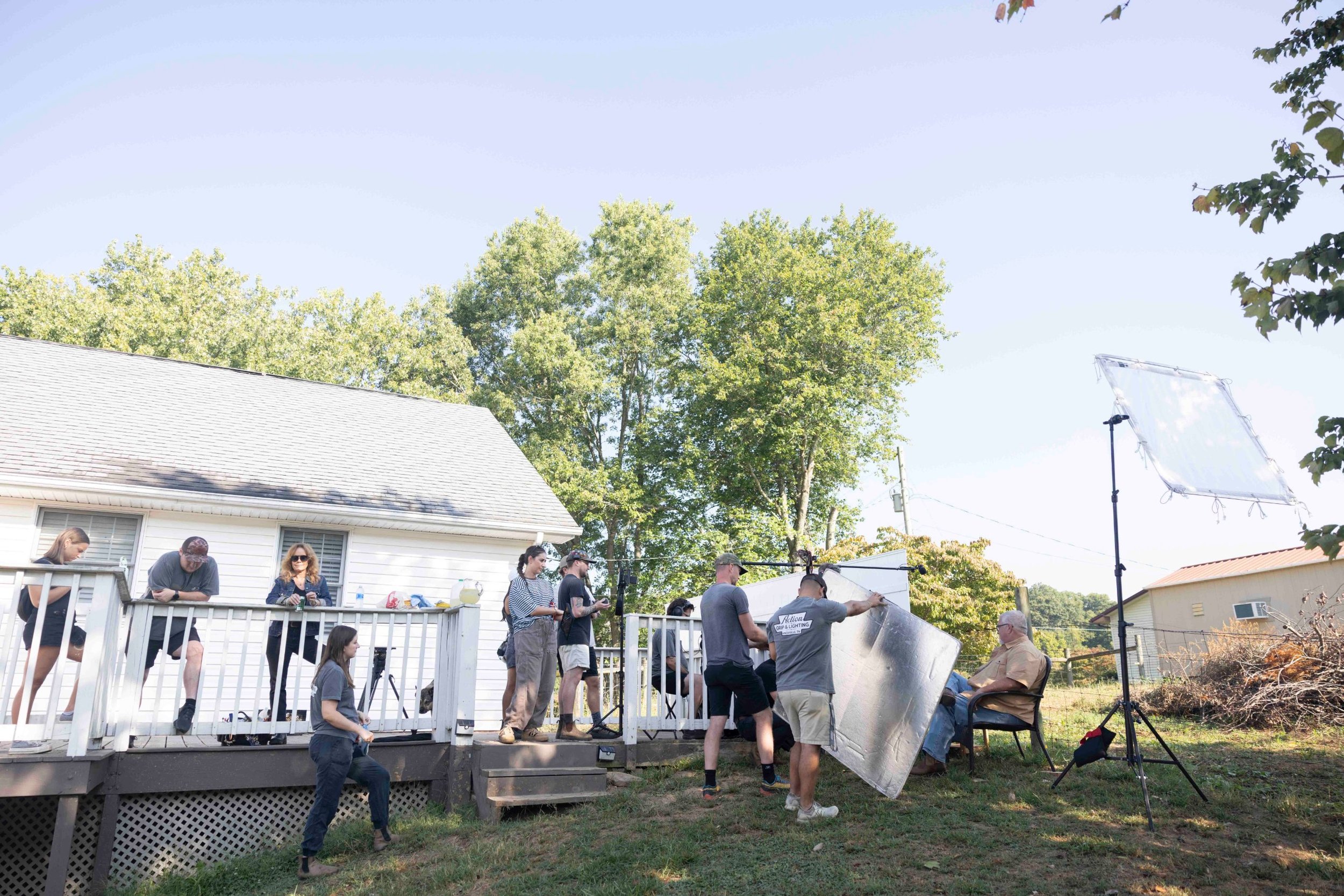 Behind the scenes of a pharma patient testimonial shoot: crew with a lighting scrim and sound boom set up beside a house while the patient sits in a chair