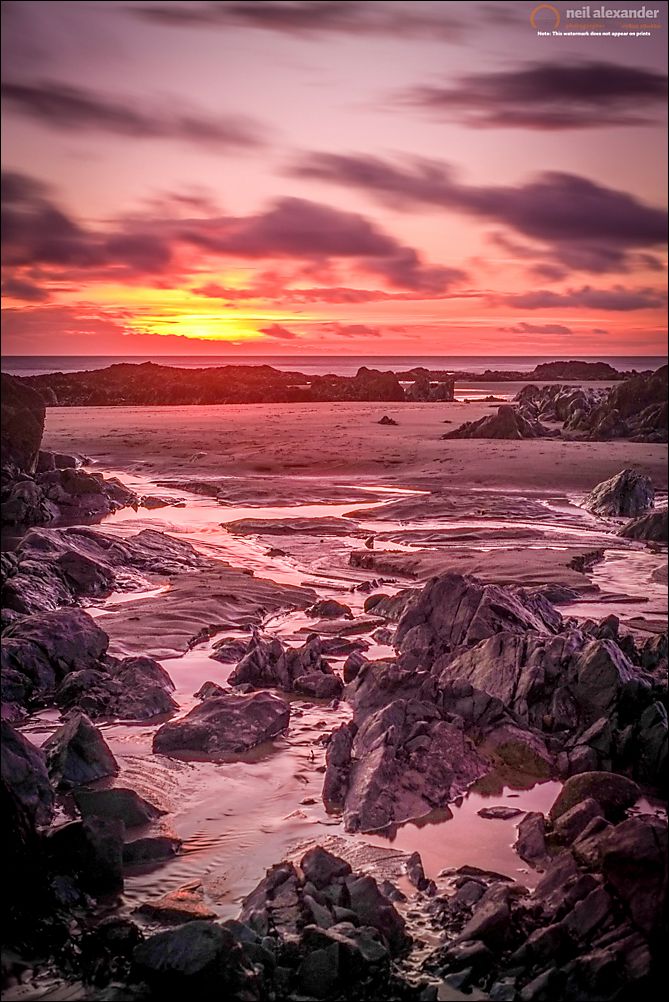 A long exposure of the sun setting over Rhosneigr beach with rocks in the foreground