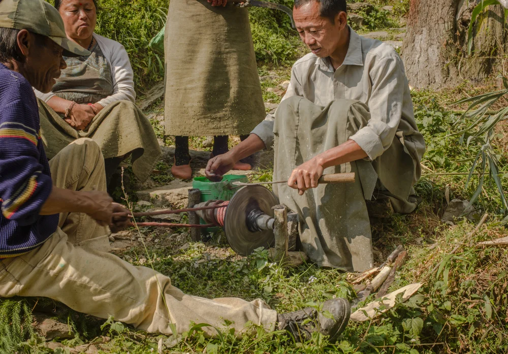Men sharpen the knives used for pruning in the tea plantation. The grinding wheel is driven by pulling back and forth on a rope wrapped around a drive spindle.
