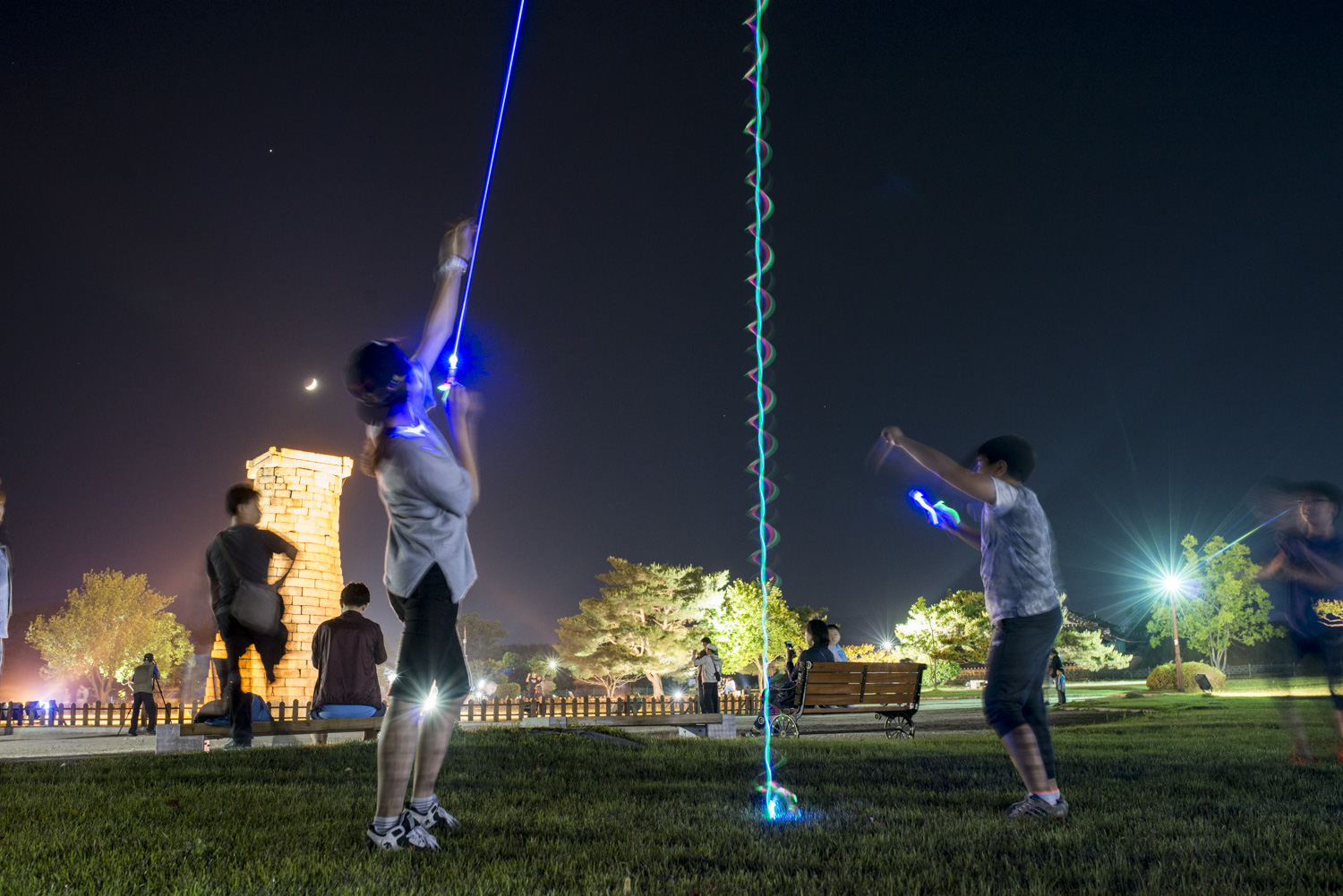 A family is having fun with some glowing helicopter toys. In the background is Cheomseongdae Observatory. Built in the 7th century A.D., it is the oldest surviving observatory in Eastern Asia. The moon is hanging out above the observatory.