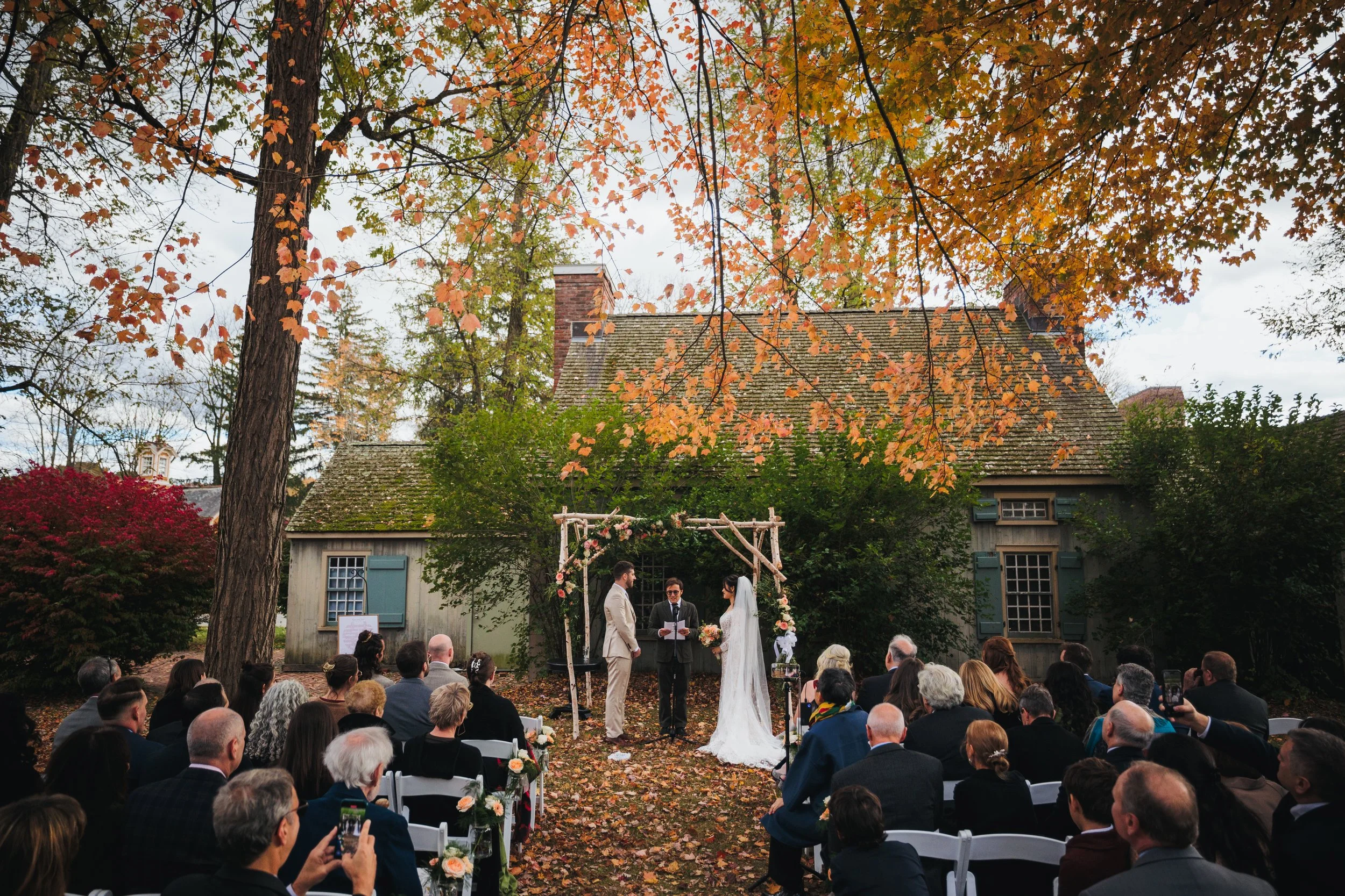 Outdoor wedding ceremony in front of a small rustic house surrounded by trees with autumn foliage. The bride and groom stand under a wooden arch with officiant. Guests seated on white chairs observe the ceremony.