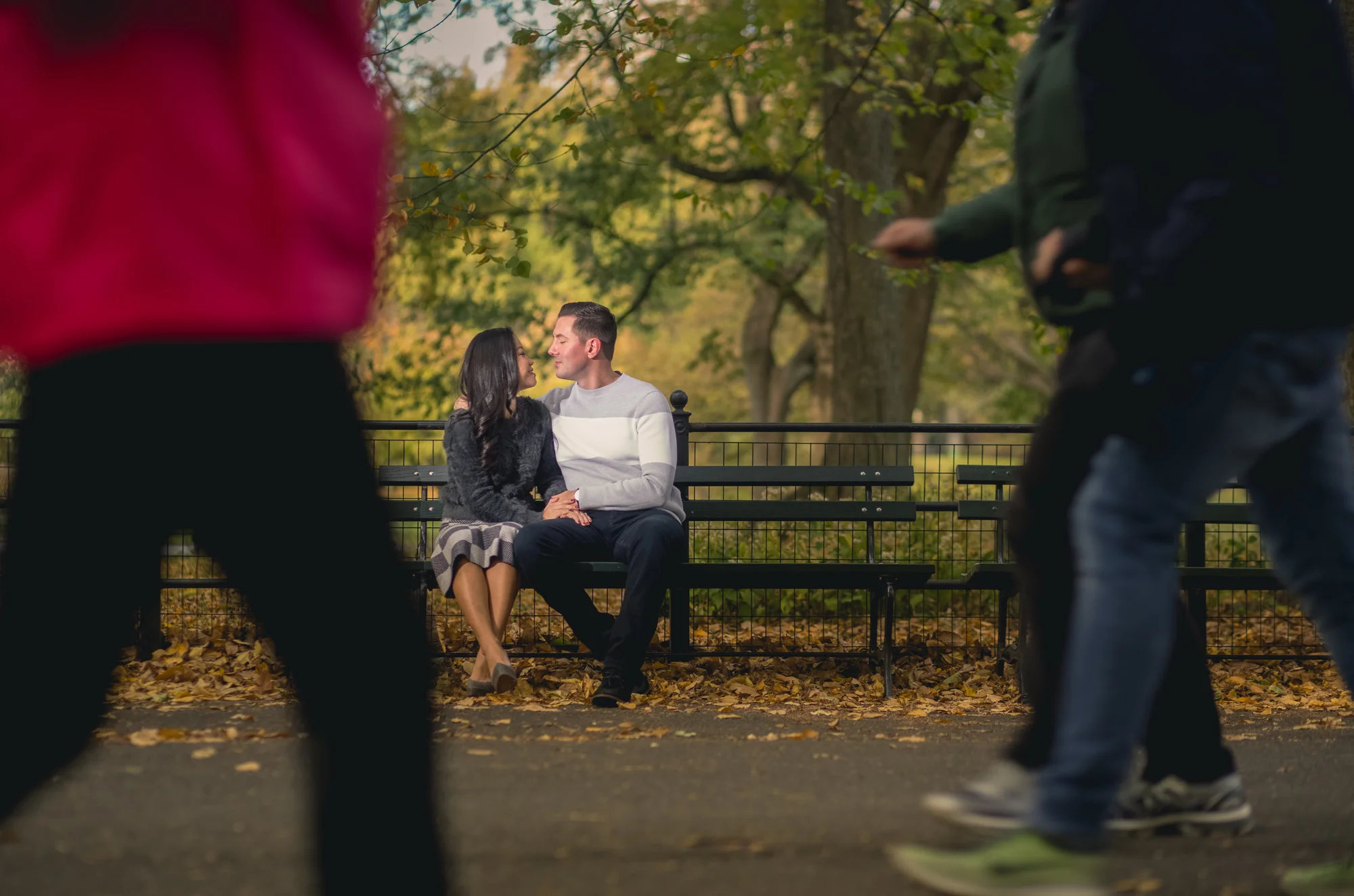 A couple sitting closely on a park bench, looking into each other's eyes, surrounded by trees with fallen leaves. Two people are walking past in the foreground, partially obscuring the view.