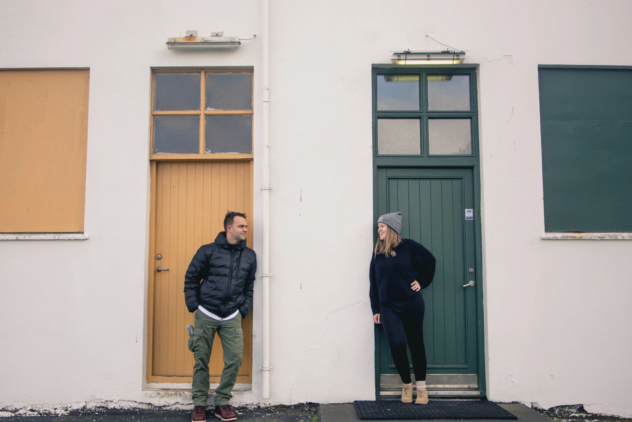 A man and woman standing outside in front of a white wall with two doors and boarded-up windows, smiling at each other.