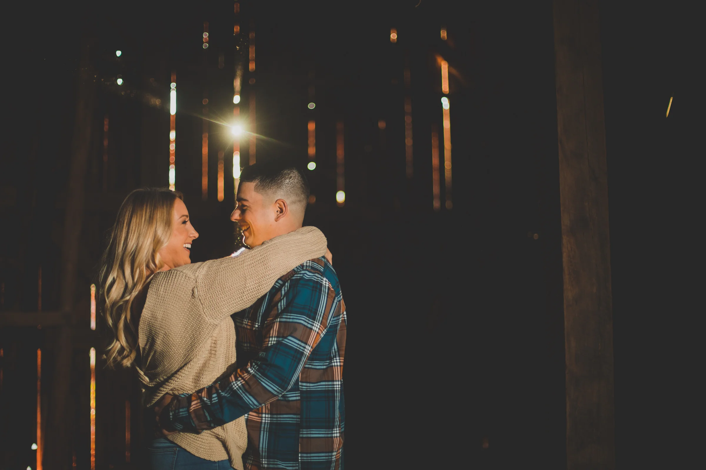 A smiling couple hugging and looking at each other in a dimly lit barn with sunlight streaming through gaps in the wooden walls.