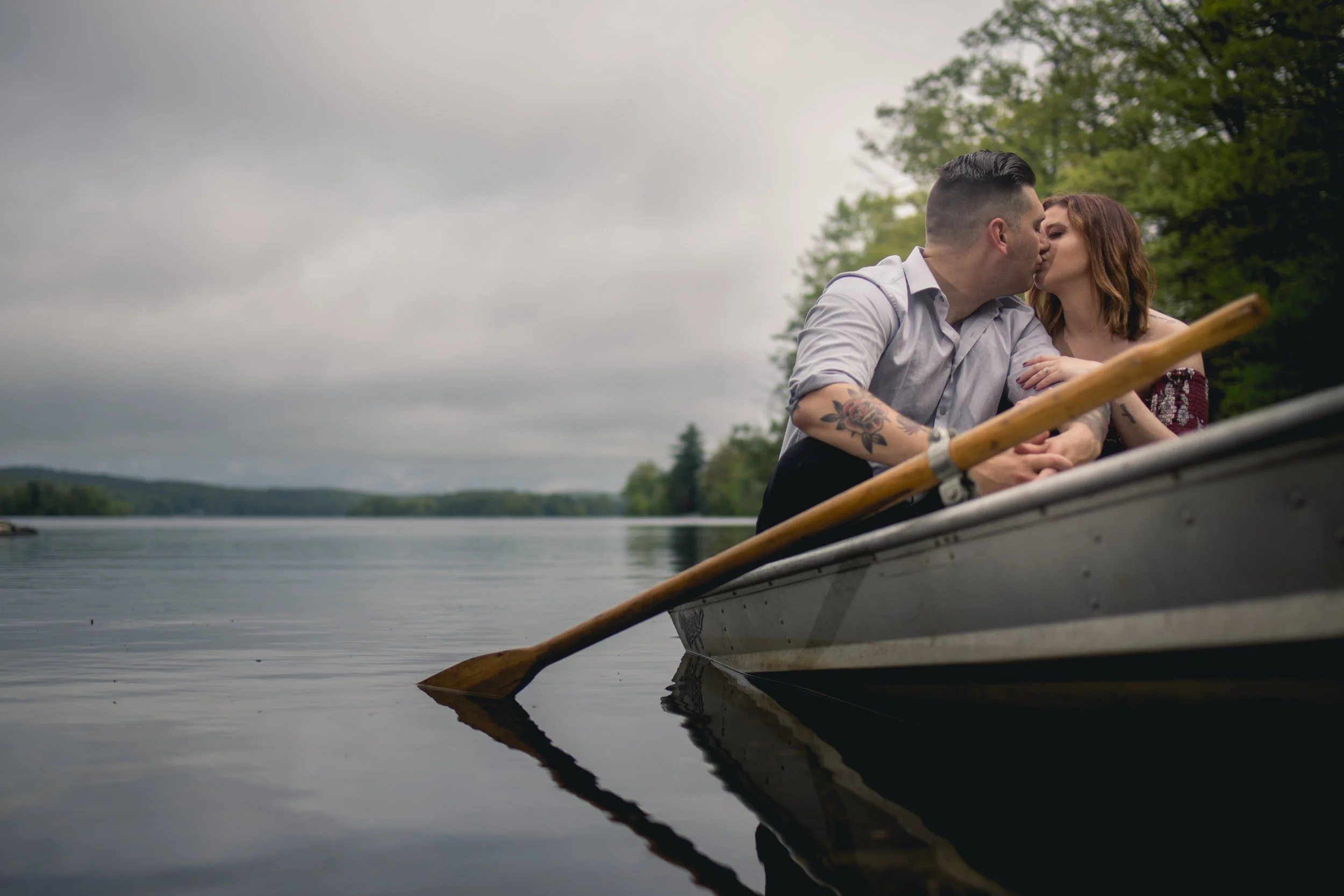 A couple sitting in a boat on calm water, sharing a kiss, with trees and cloudy sky in the background.