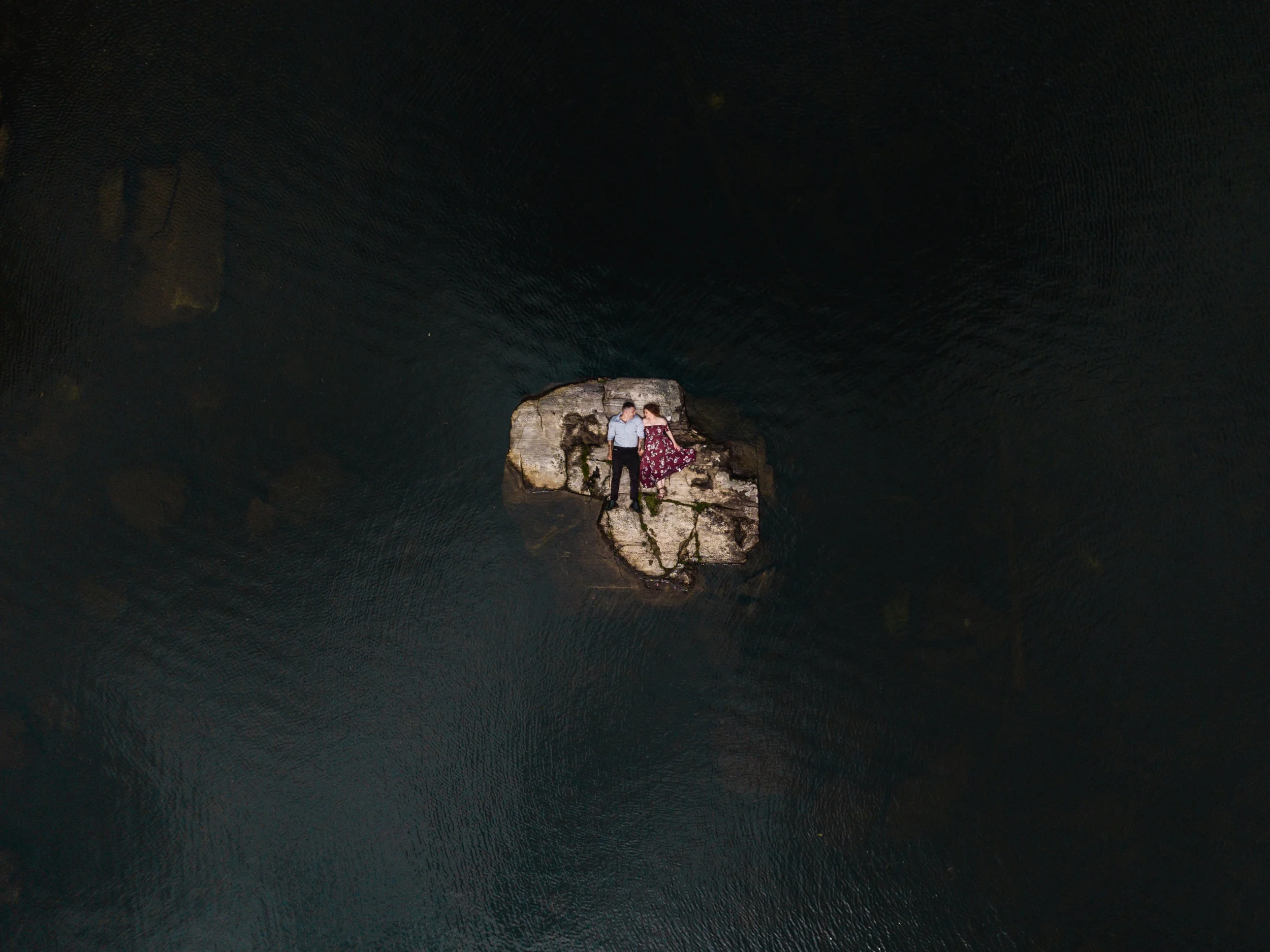 A couple lying on a large rock in a body of water, holding hands and looking at each other.