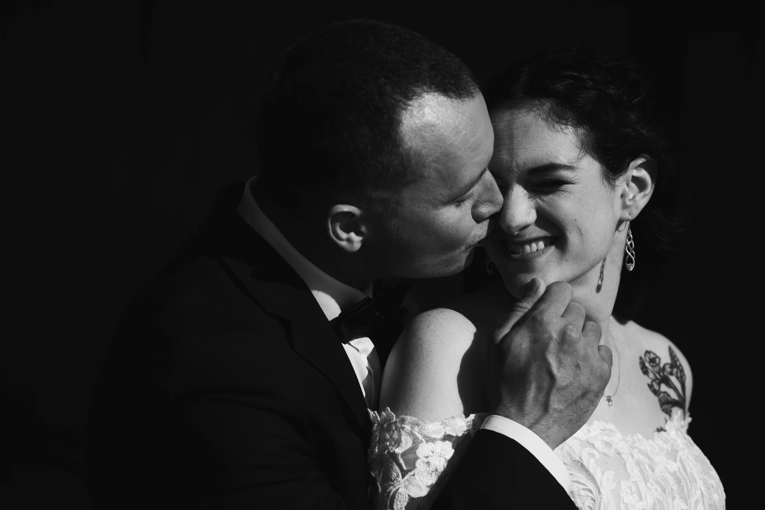 A black and white photo of a couple on their wedding day, with the groom kissing the bride on the cheek.