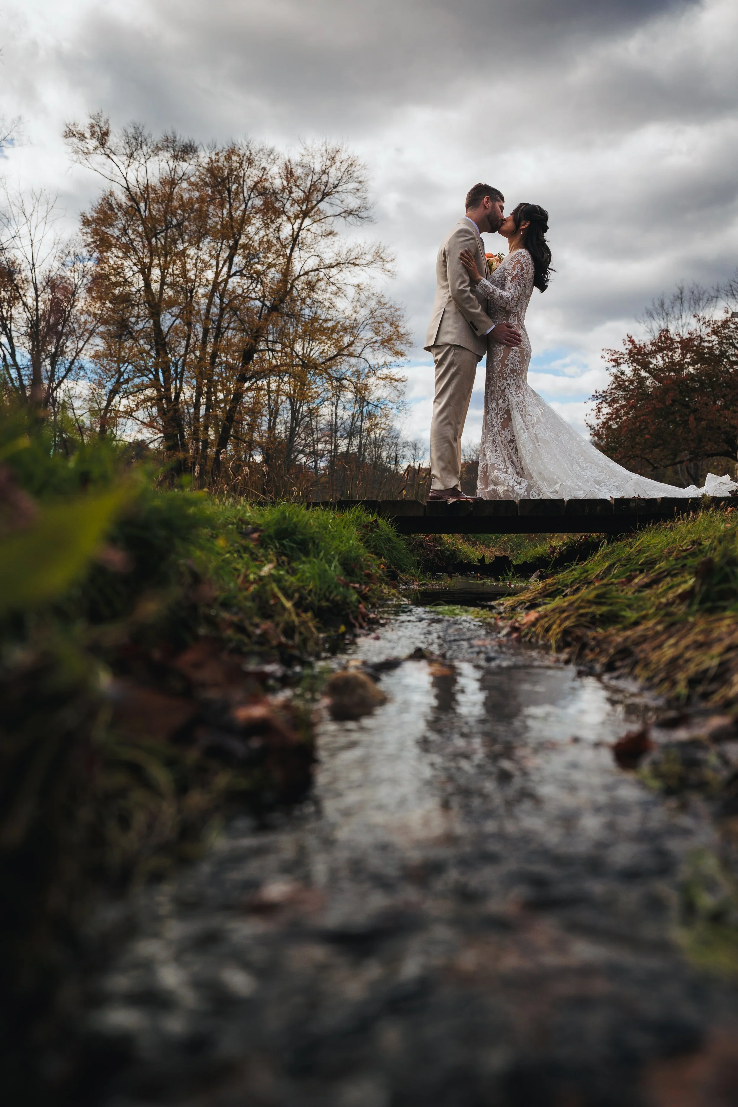 A newlywed couple sharing a kiss on a bridge over a stream in fall.