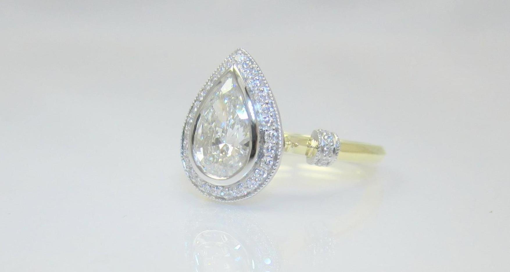  A two-tone engagement ring handcrafted by Booth Custom Jewelers in the belle époque style, featuring 18k yellow gold and platinum. The pear-shaped diamond is set in a platinum bezel and is surrounded by a bead-set diamond halo. Platinum finials on t