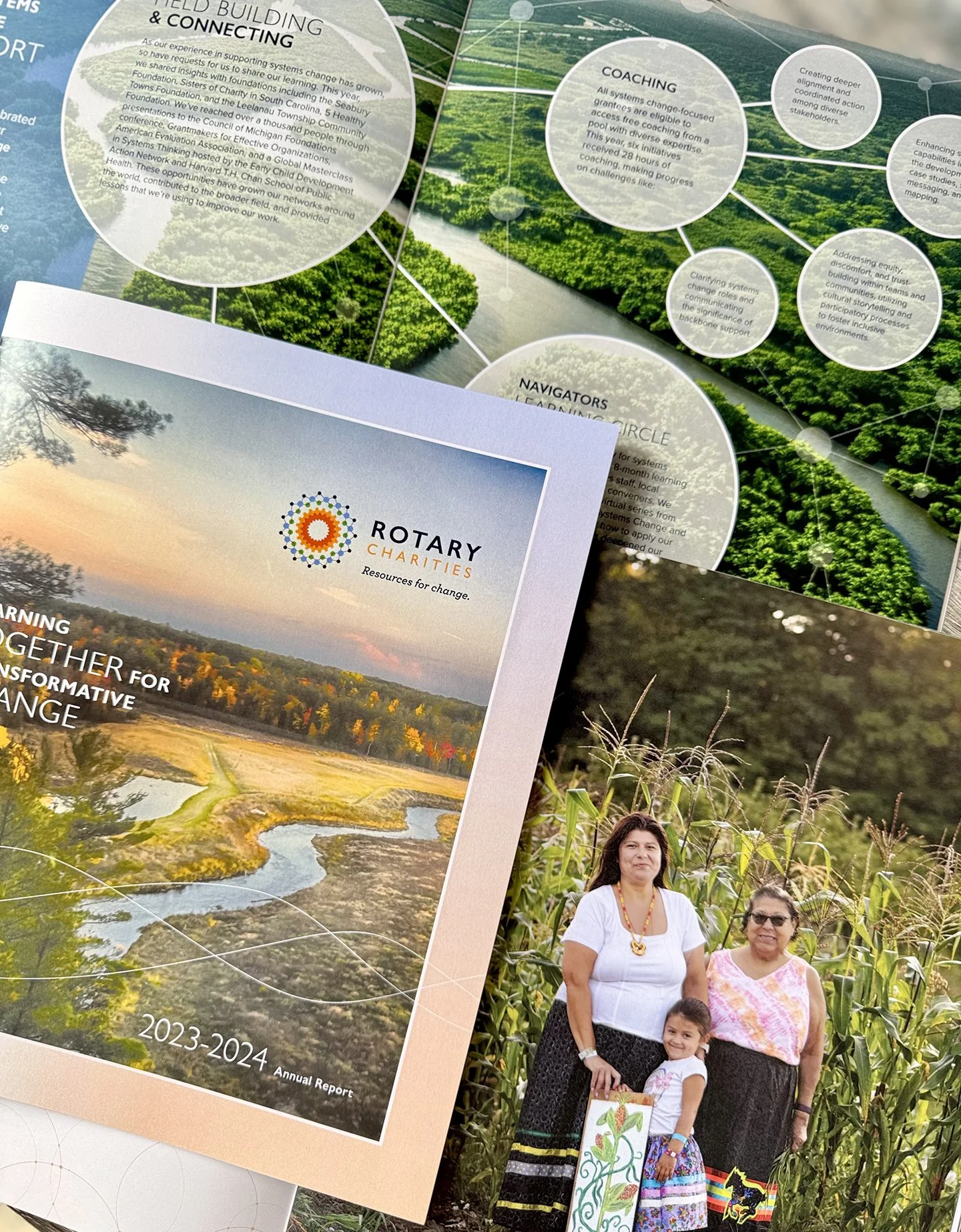 A photo showing various documents, including a Rotary Charities annual report cover featuring a sunset landscape with trees and a pond, and a photograph of two women and a young girl standing in a cornfield, smiling at the camera.