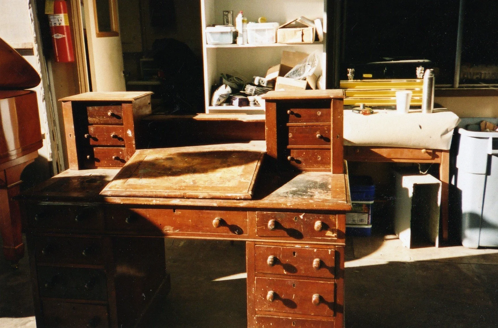 Old wooden desk with multiple drawers, some drawers are missing or damaged, in a cluttered workshop or garage area with various tools and supplies on shelves in the background.