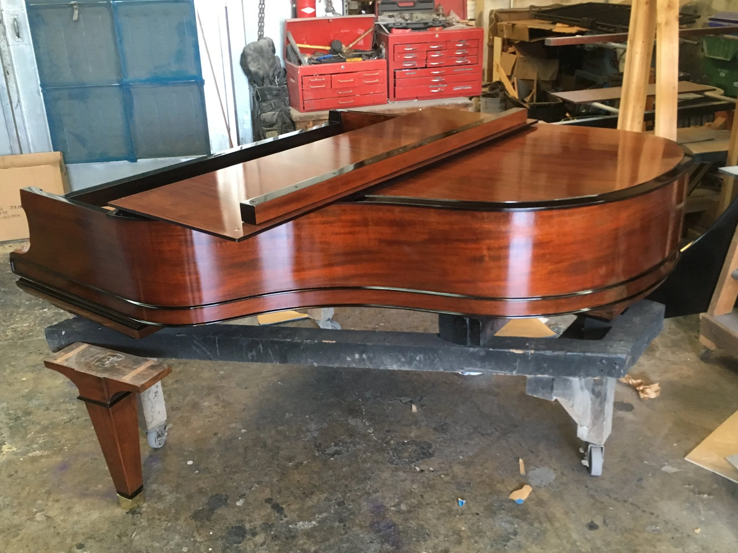 A polished wooden grand piano with a closed lid in a workshop, surrounded by tools and shelves.