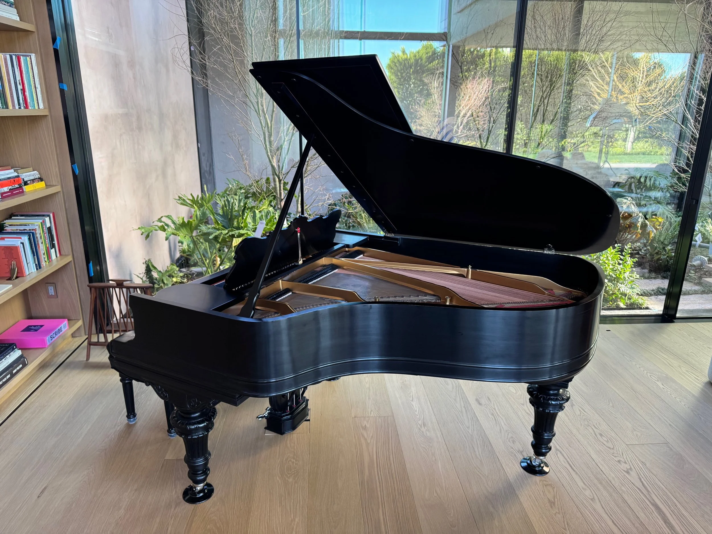 A black grand piano with an open lid, situated in front of large glass windows with a garden view. To the left, there is a bookshelf filled with books and a small pink box.