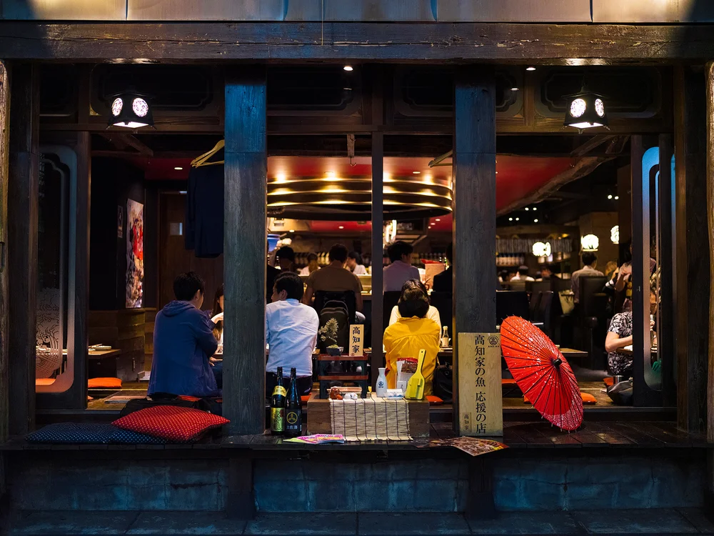 A nighttime photograph of a Tokyo bar storefront with large windows framing the interior scene.