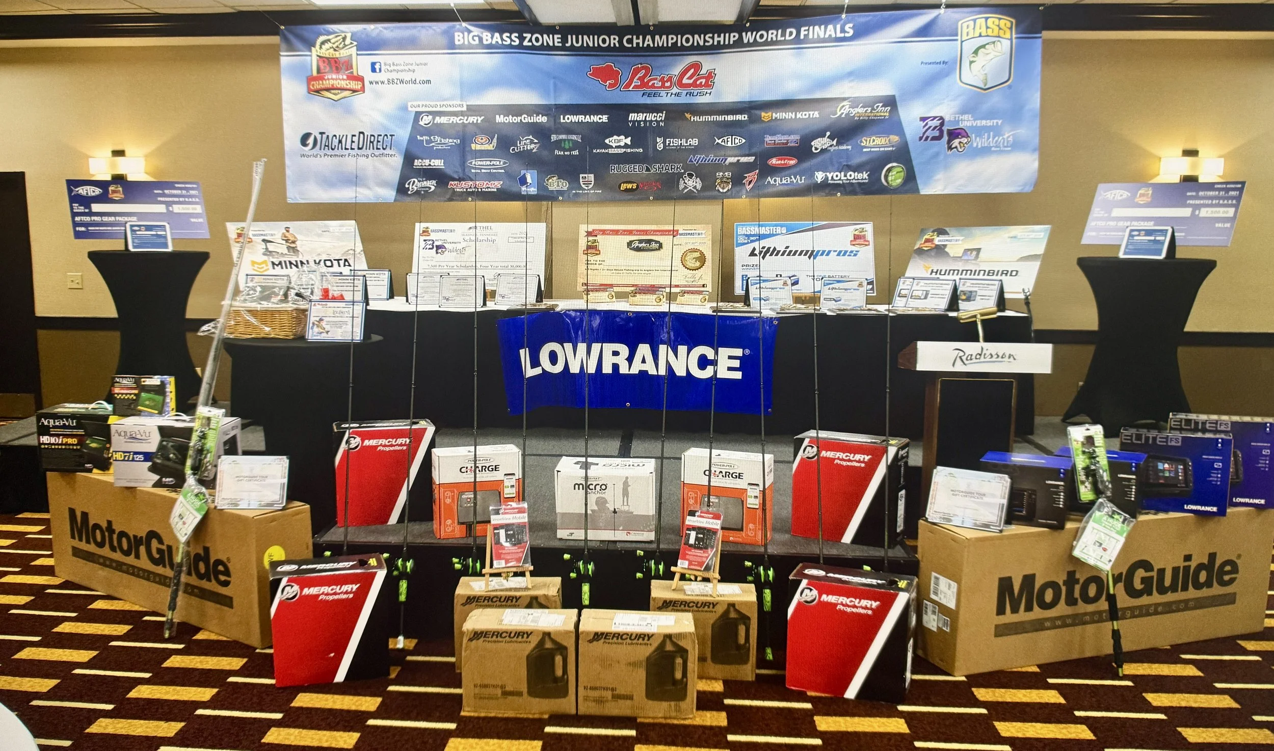 Exhibit table at the Big Bass Zone Junior Championship World Finals displaying fishing gear and equipment from brands like Lowrance, Mercury, and MotorGuide, with a large banner overhead and various prizes and products arranged around the table.
