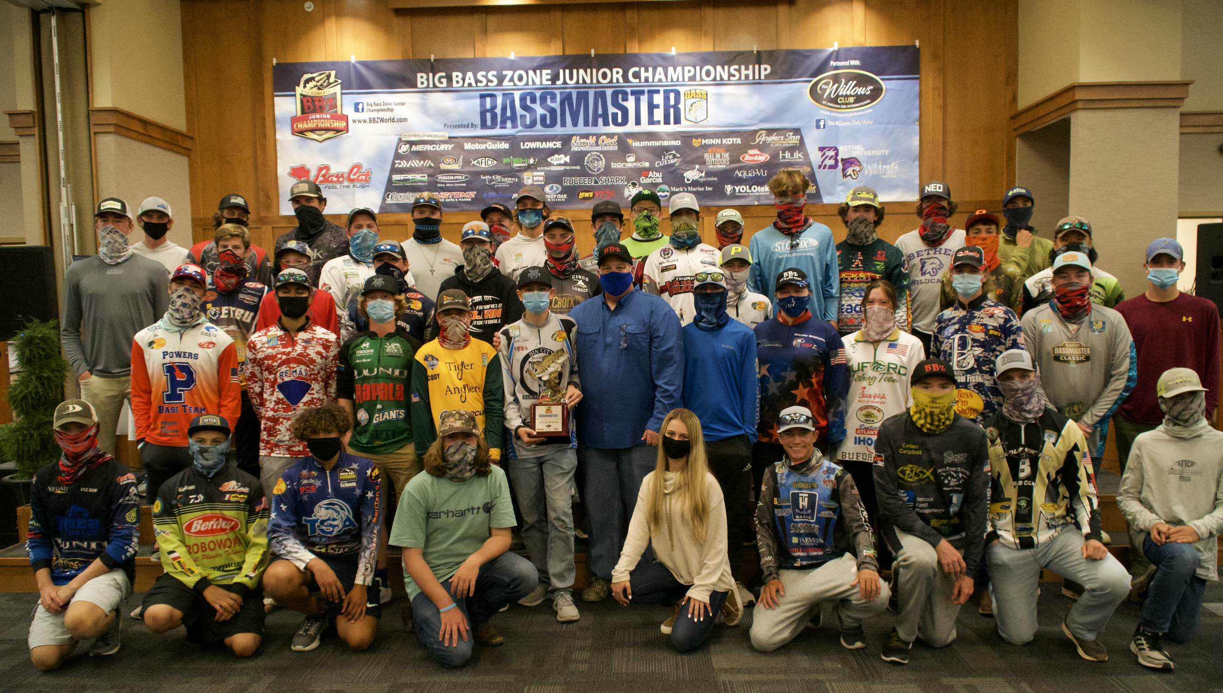 Group of young racers and adults in a large room with a wooden wall background, standing in front of a banner that reads "Big Bass Zone Junior Championship" and "Bassmaster," posing for a photo, many wearing colorful race jerseys and masks.