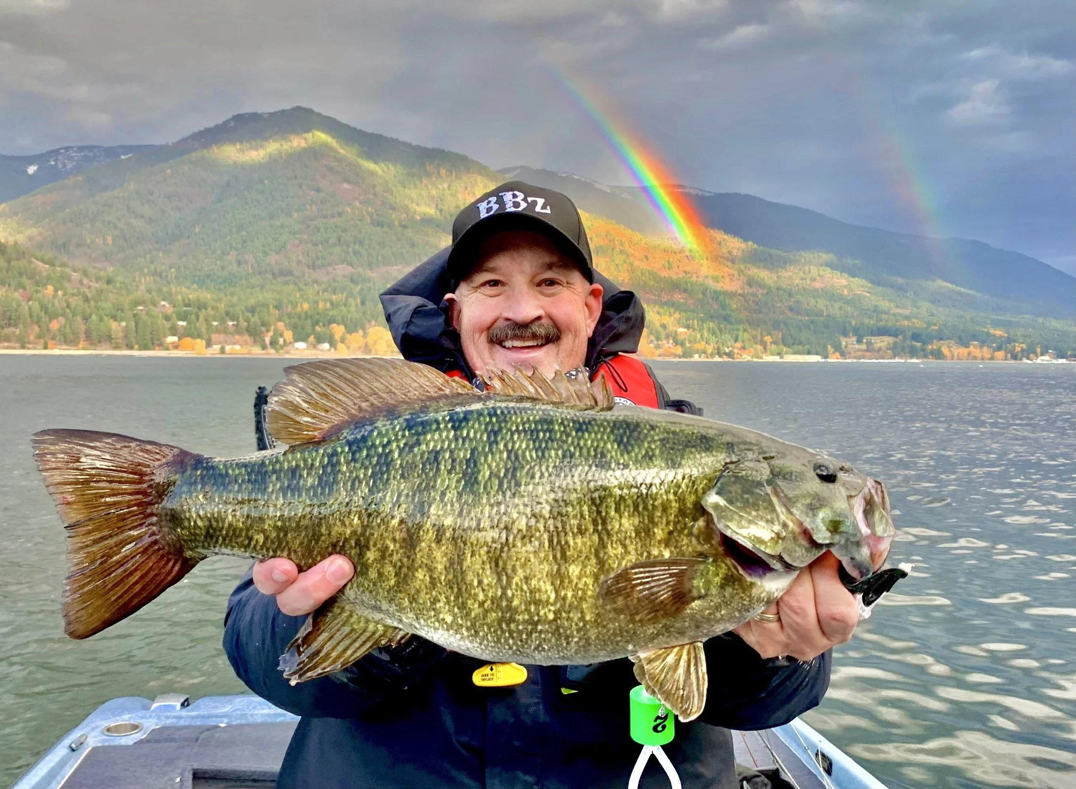 A man smiling and holding a large fish, with a colorful rainbow, mountain, and lake in the background.