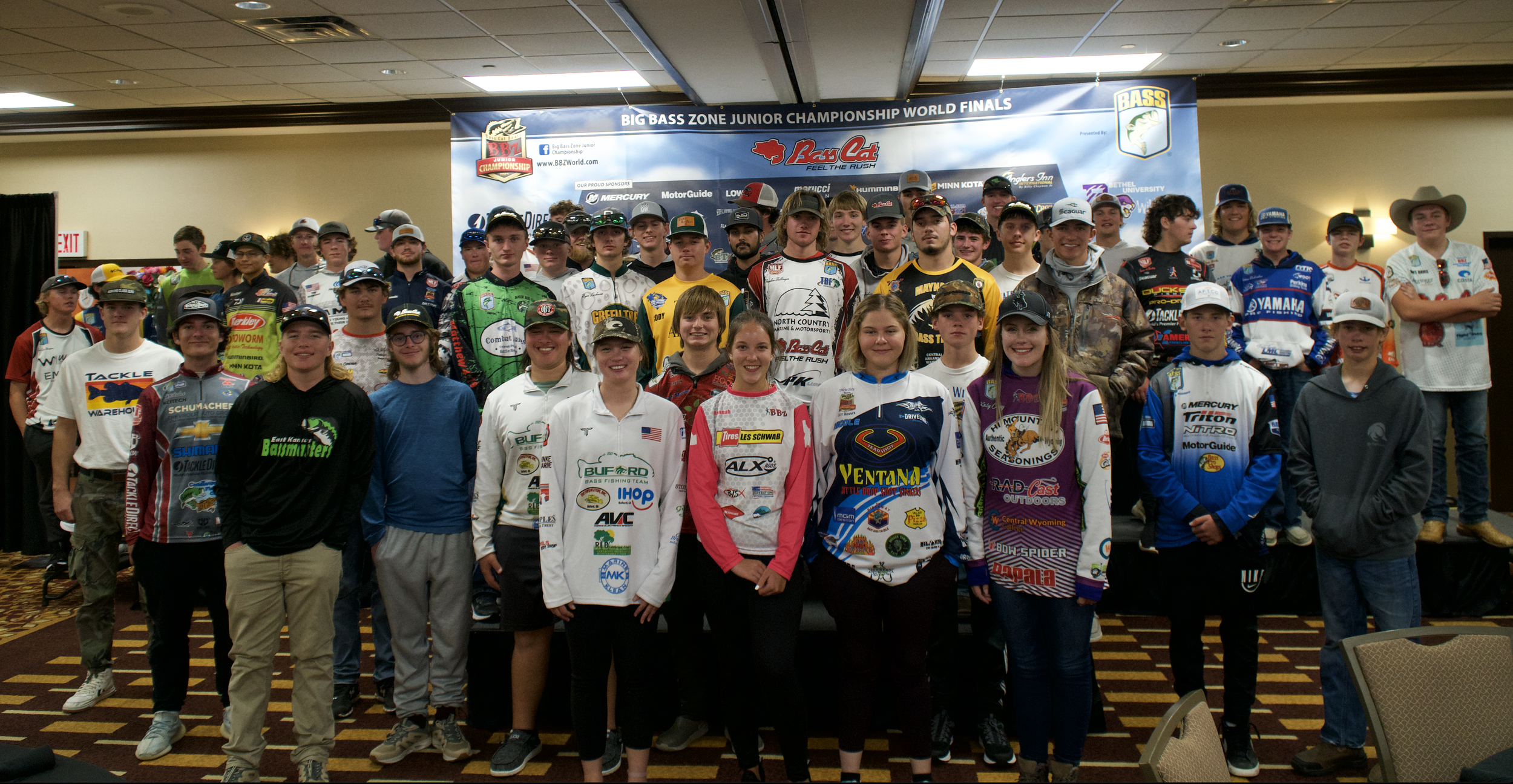 Group of young individuals, mostly teenagers, gathered on a stage for a championship event, with a banner above reading 'Big Bass Zone Junior Championship World Finals'. They are dressed in various sports jerseys and casual clothing, some wearing hats, and are smiling for the photo.