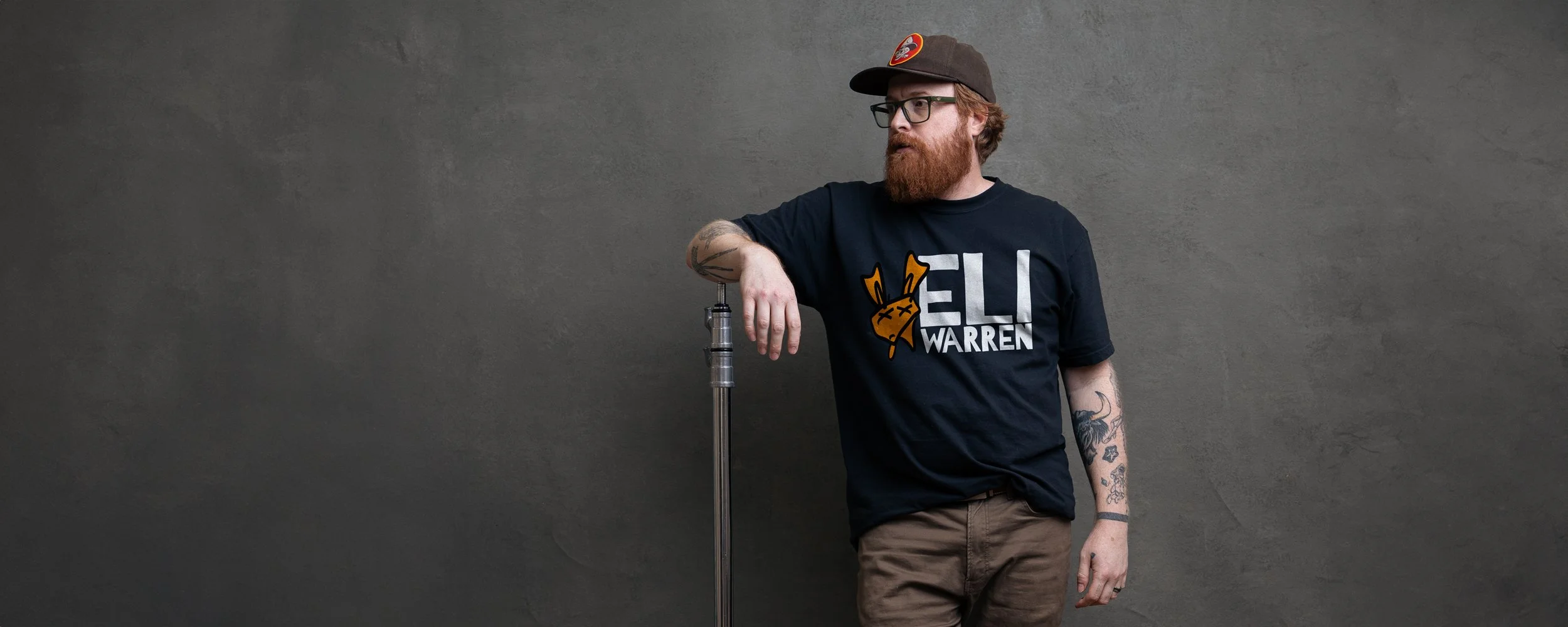 Picture of Eli Warren wearing his logo shirt in front of a gray painted backdrop