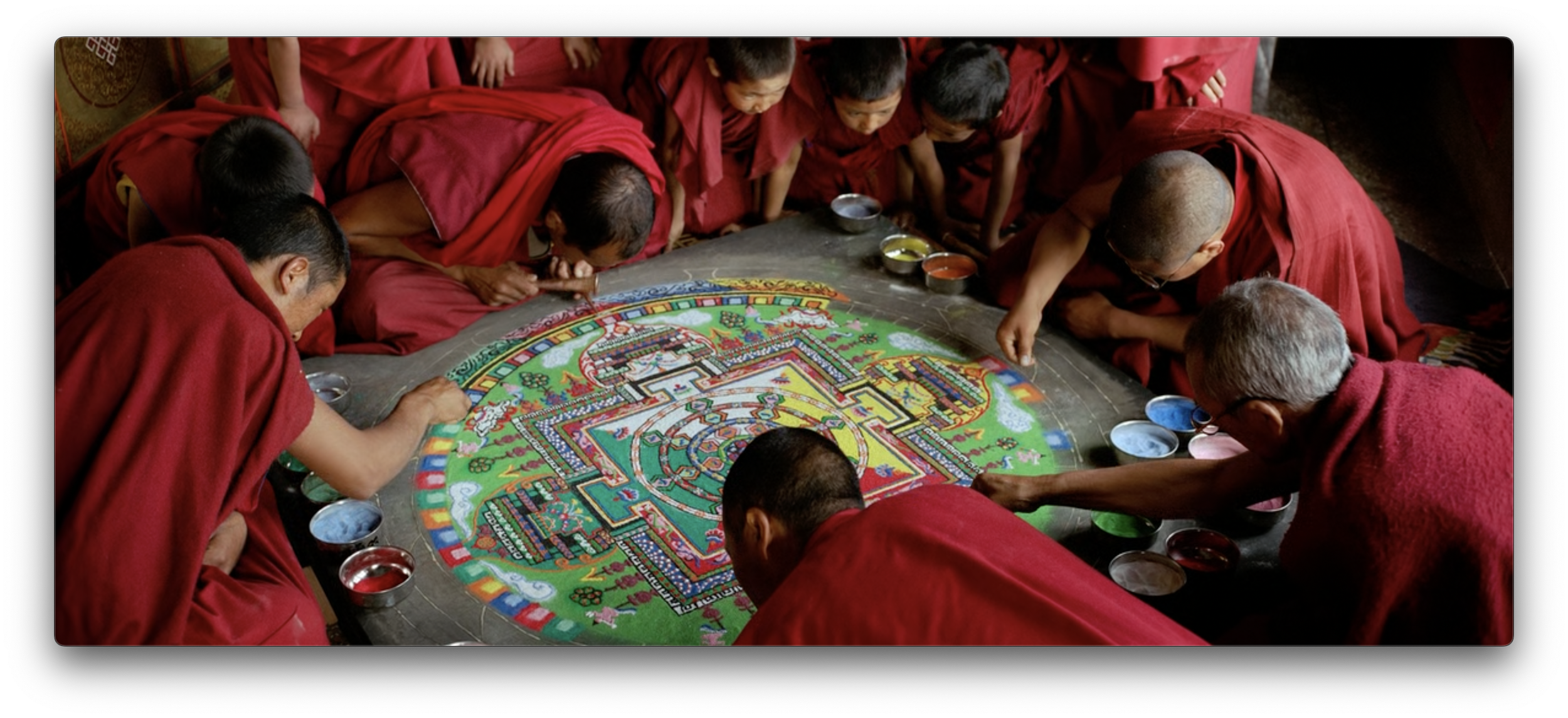 Tibetan monks in red robes work around a circular mandala with various colors of sand
