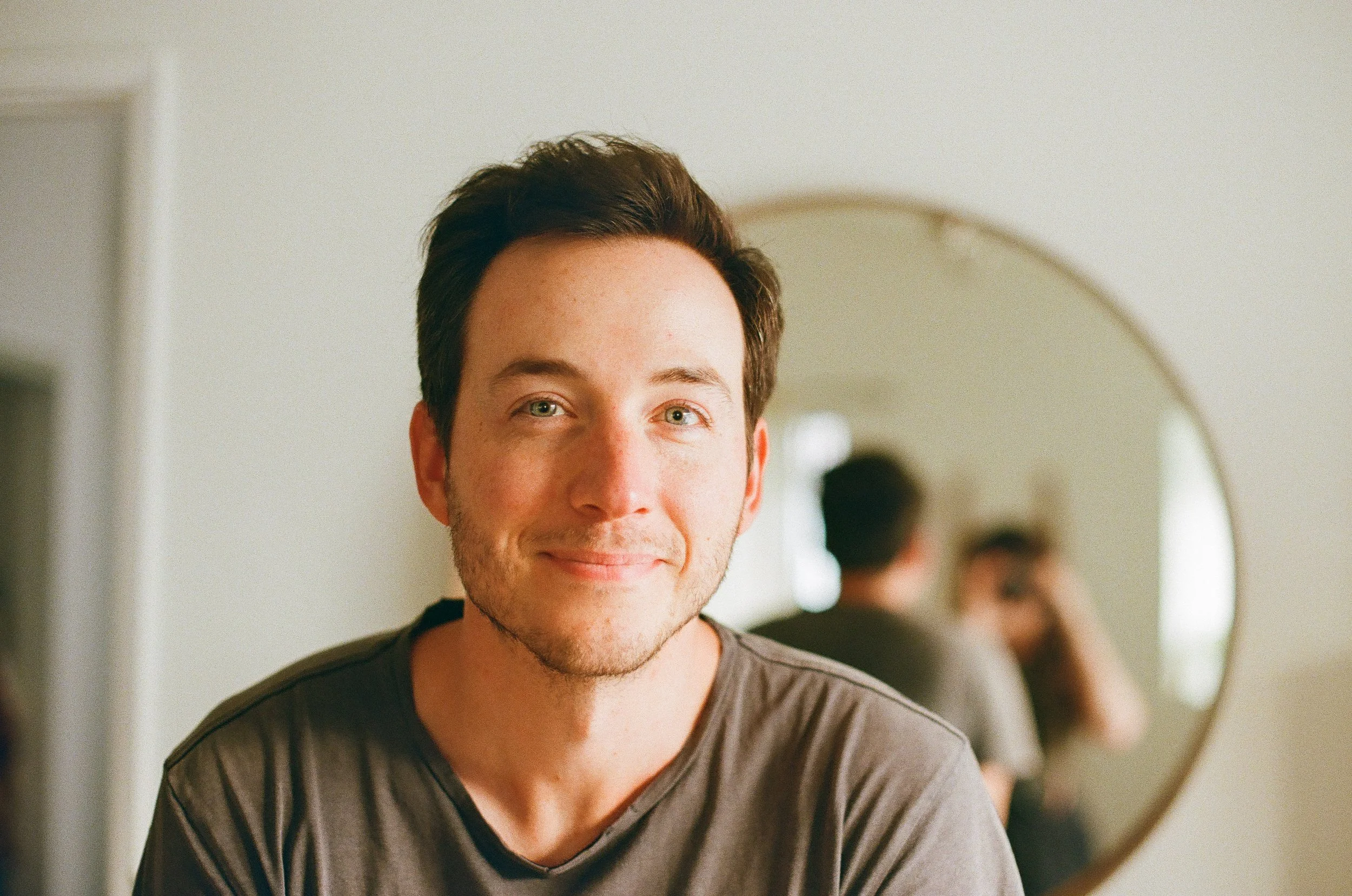 Portrait of a young man with short dark hair and green eyes, smiling at the camera, standing in front of a mirror with a blurred background and a person visible in the reflection.