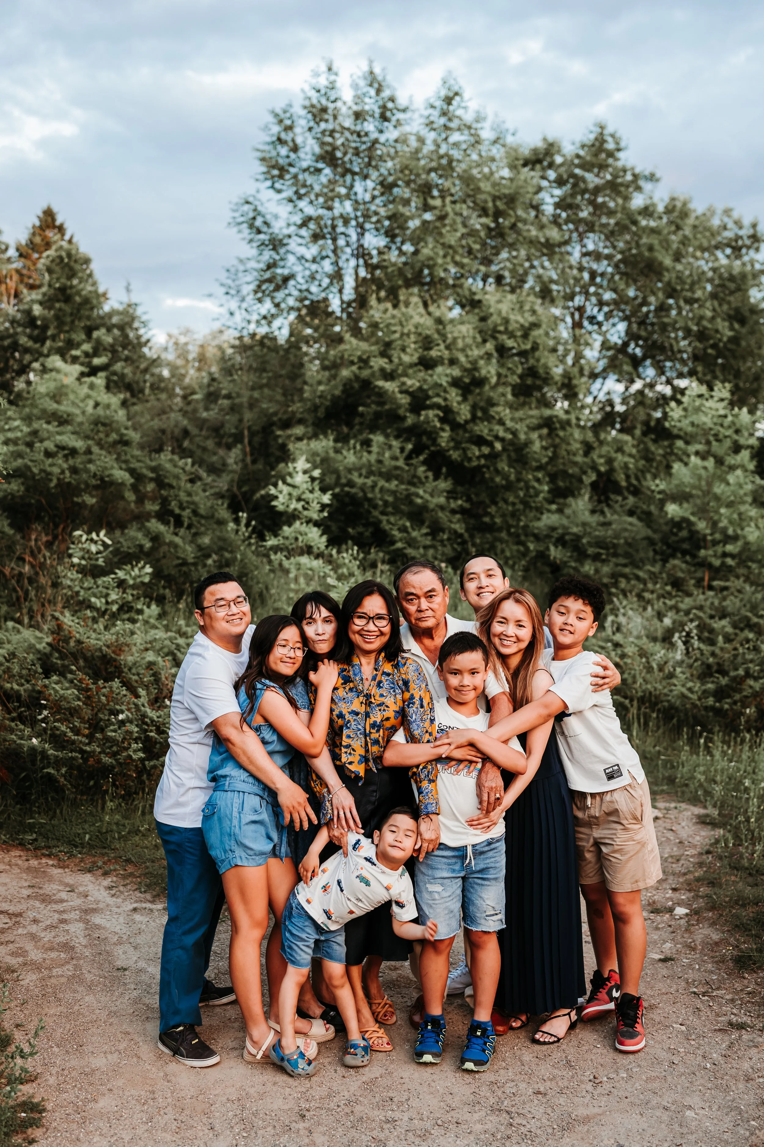 A large family of eleven people, including children and adults, standing together outdoors on a dirt path with green trees and a partly cloudy sky in the background, smiling and embracing each other.