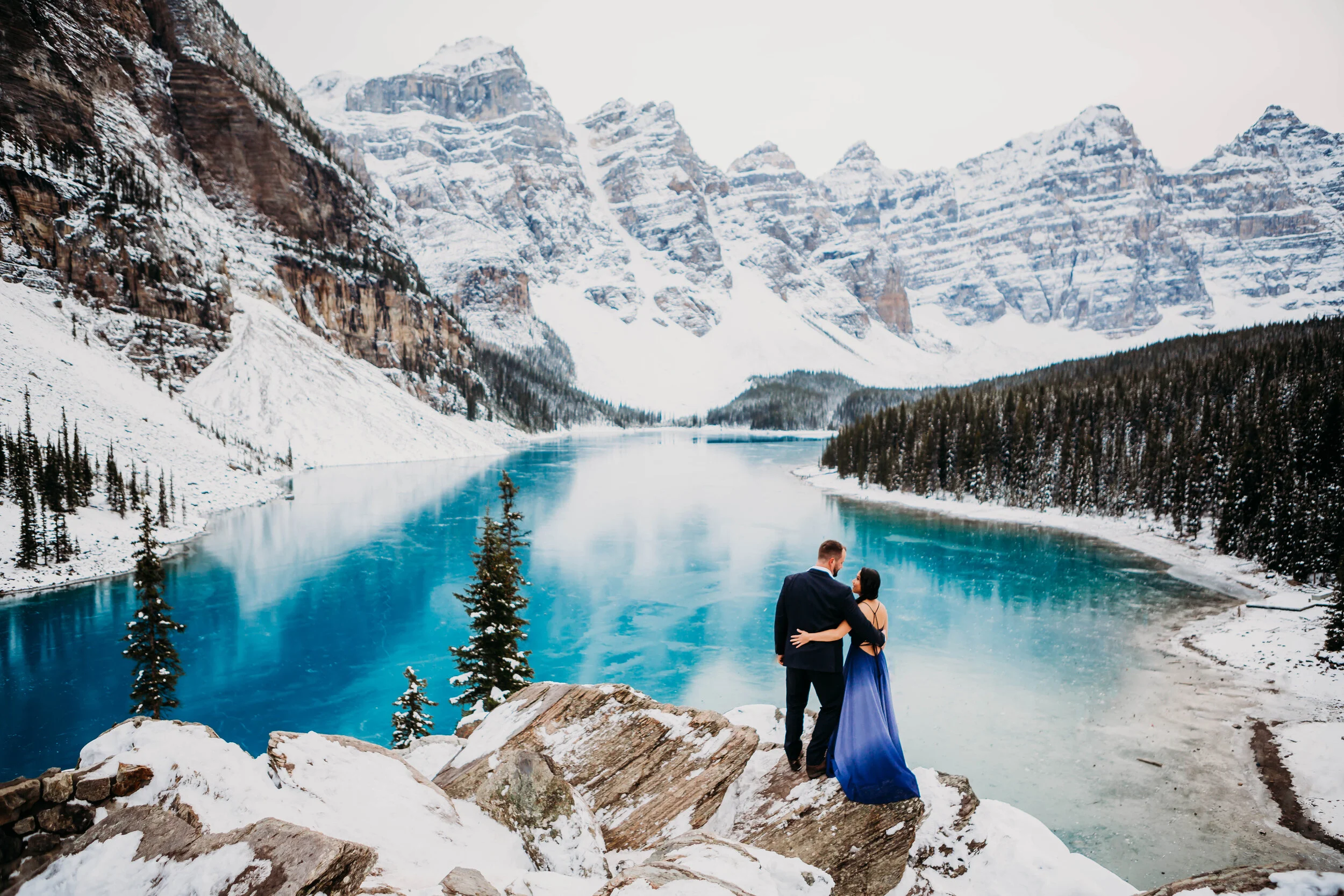 A couple in formal attire stands on a rocky snowy ledge overlooking a partially frozen lake surrounded by snow-covered mountains and pine trees.