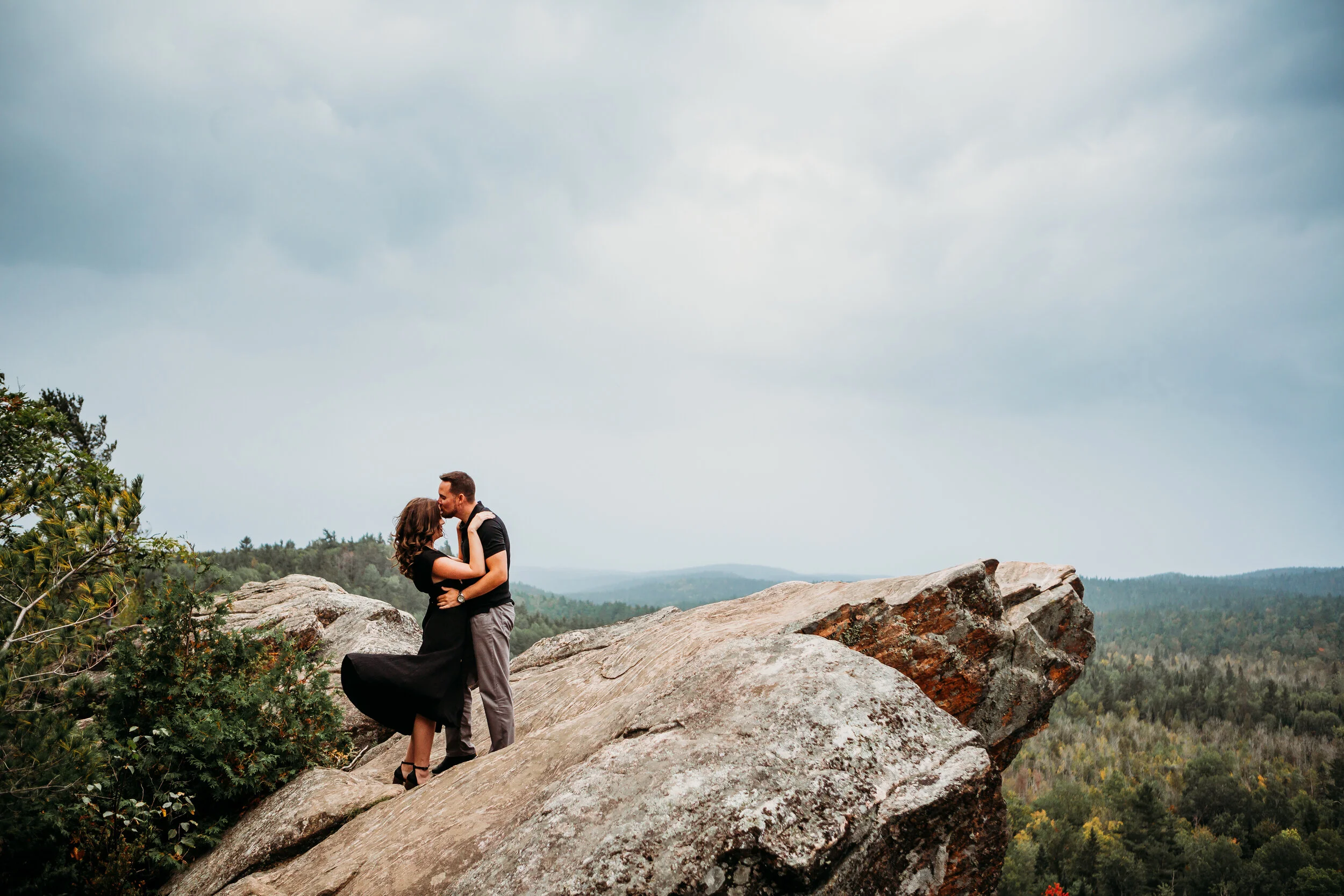 A couple embraces and kisses on a large rock formation overlooking a forested landscape under cloudy sky.