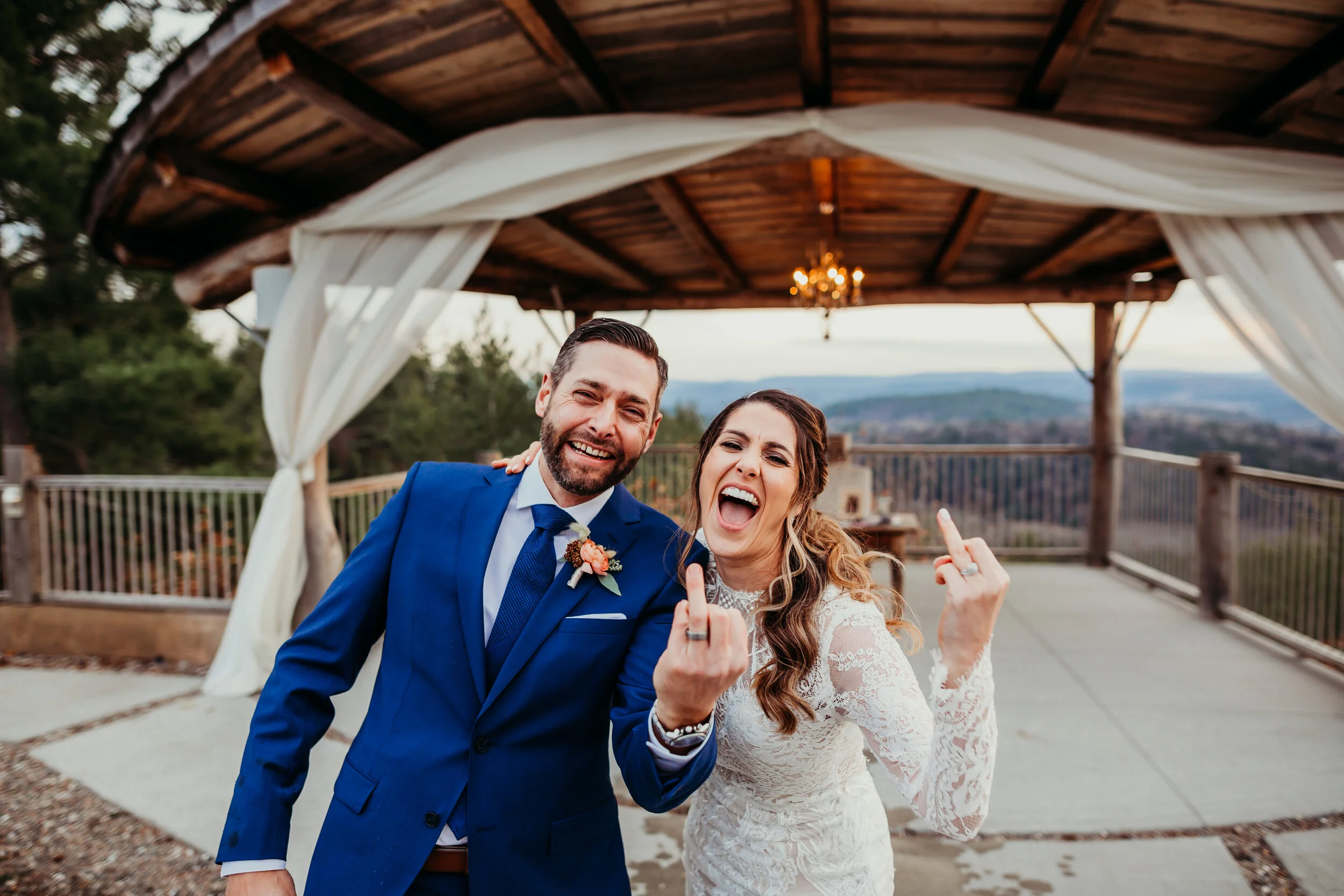 A newlywed couple, dressed in wedding attire, making rude gestures while smiling outdoors under a wooden pavilion with white drapery.