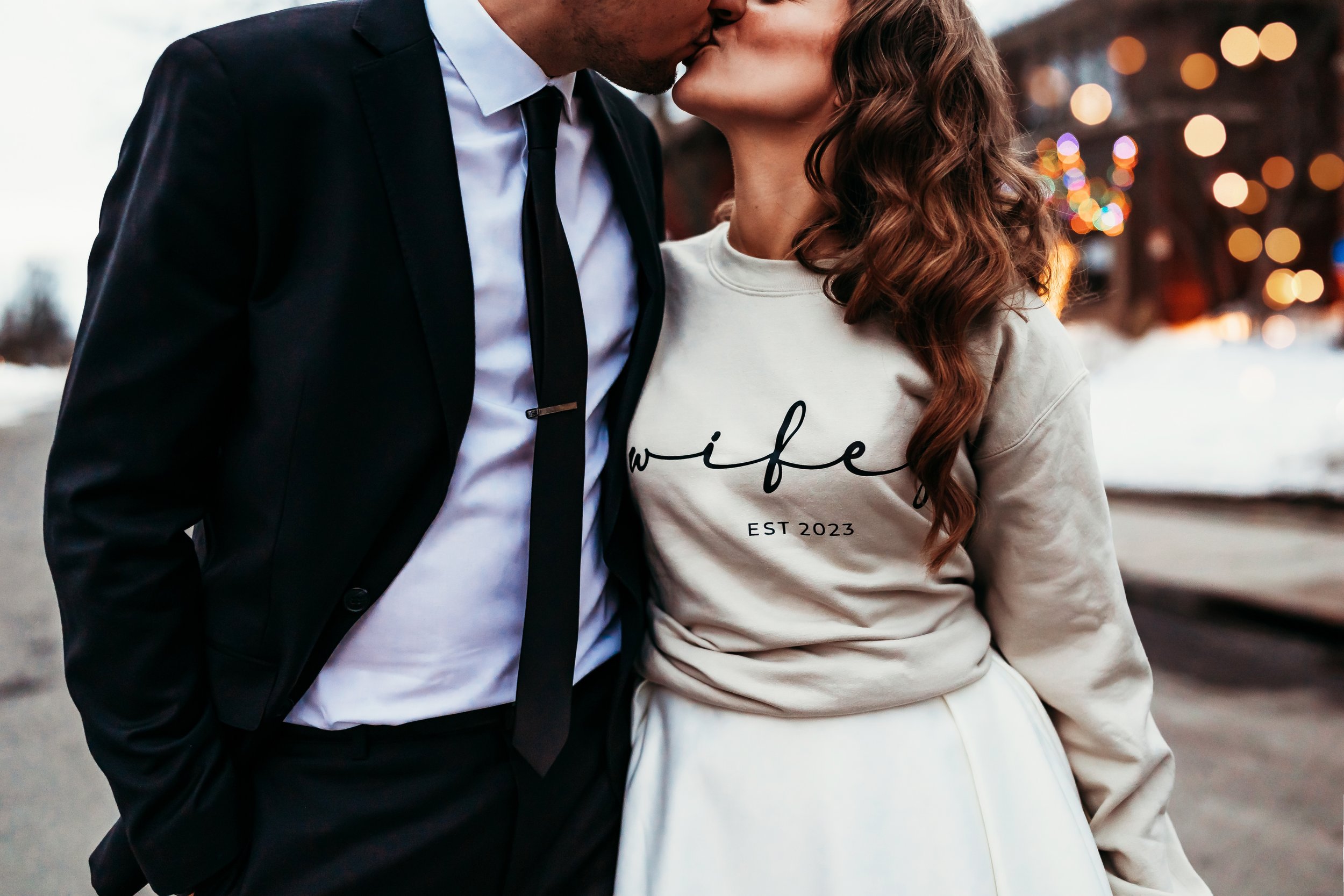 A couple sharing a kiss outdoors with blurred festive lights in the background, dressed casually and formally.