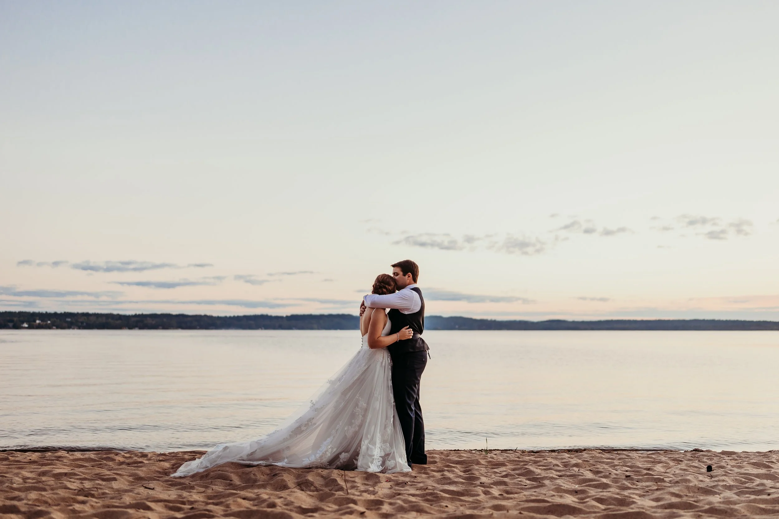 A bride and groom hugging on the sandy beach near the water, with a serene lake and cloudy sky in the background during sunset.