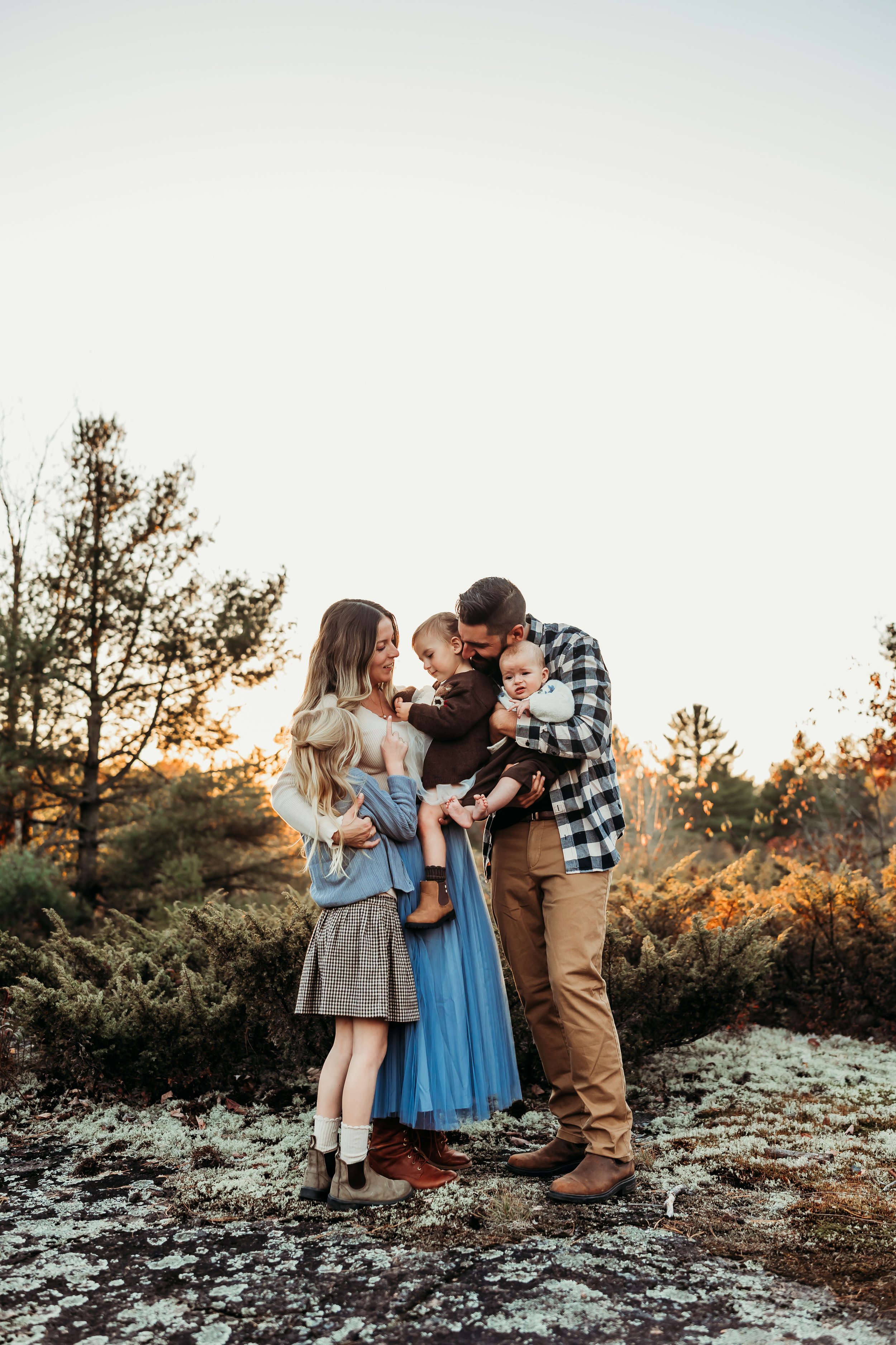 Family of five standing outdoors in fall, smiling and hugging each other, with trees and sunset in background.