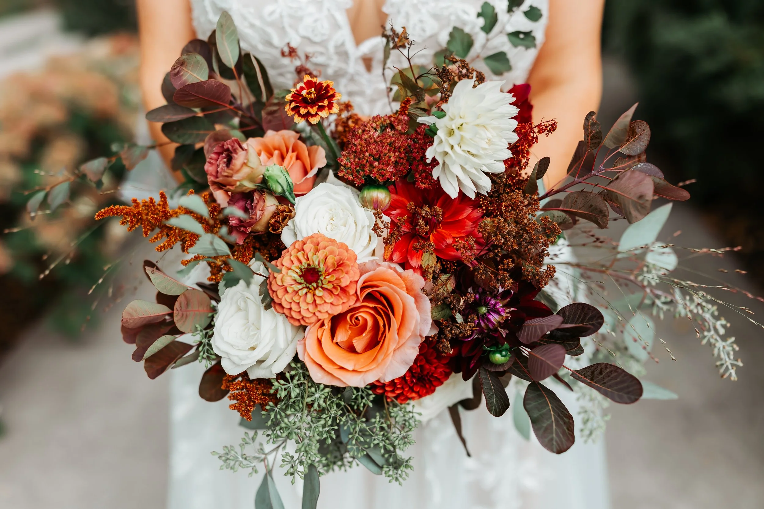 A woman holding a colorful bouquet of various flowers including orange roses, white roses, red dahlias, and other blooms, with dark and green leaves.