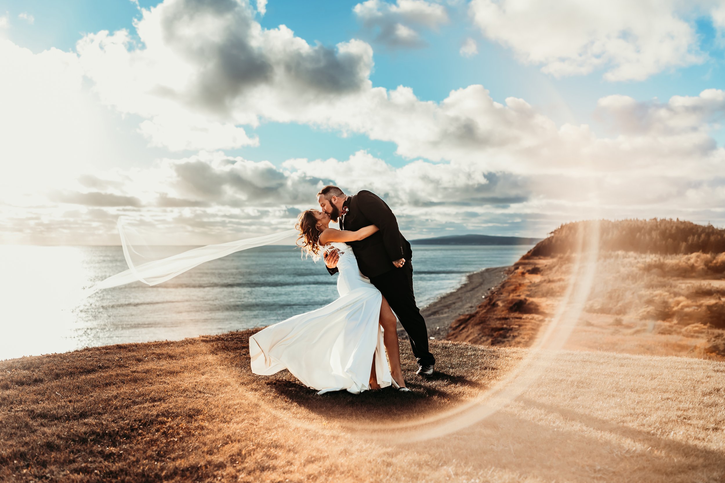 A couple in wedding attire sharing a kiss on a grassy cliffside overlooking the ocean at sunset, with a rainbow and clouds in the sky.