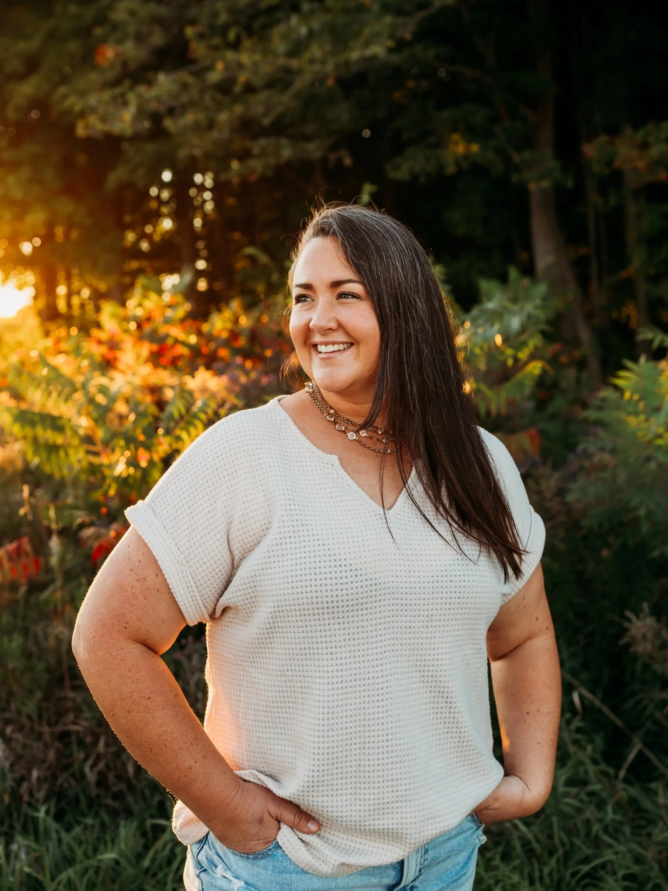 A smiling woman standing outdoors during sunset with trees and colorful foliage in the background, wearing a white short-sleeve shirt and layered necklaces.